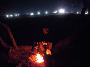 Will Kindler around the campfire at Oceti Sakowin the night of the water cannons, in the background the DAPL light towers. (Courtesy of Will Kindler)