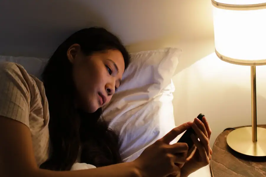 Mulher deitada na cama à noite usando o celular sob a luz de um abajur Mulher deitada na cama à noite usando o celular sob a luz de um abajur