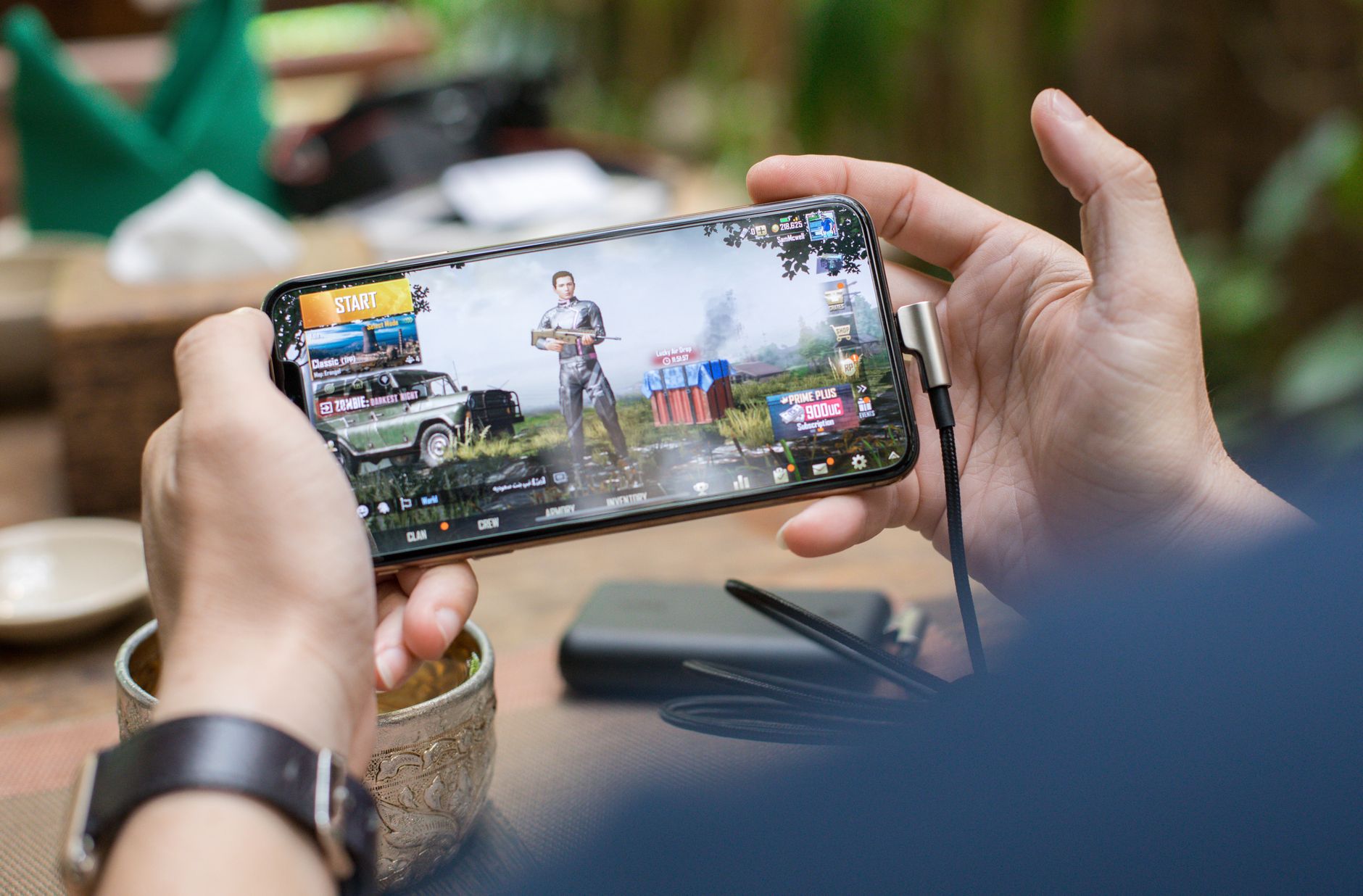 Celular para jogos: o que realmente importa Person playing PUBG on smartphone outdoors with charging cable attached.