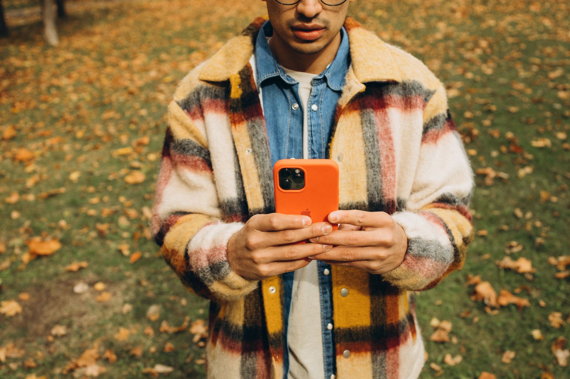 Como melhorar o sinal do celular em casa A person in a plaid jacket holds a smartphone in a park covered with autumn leaves.