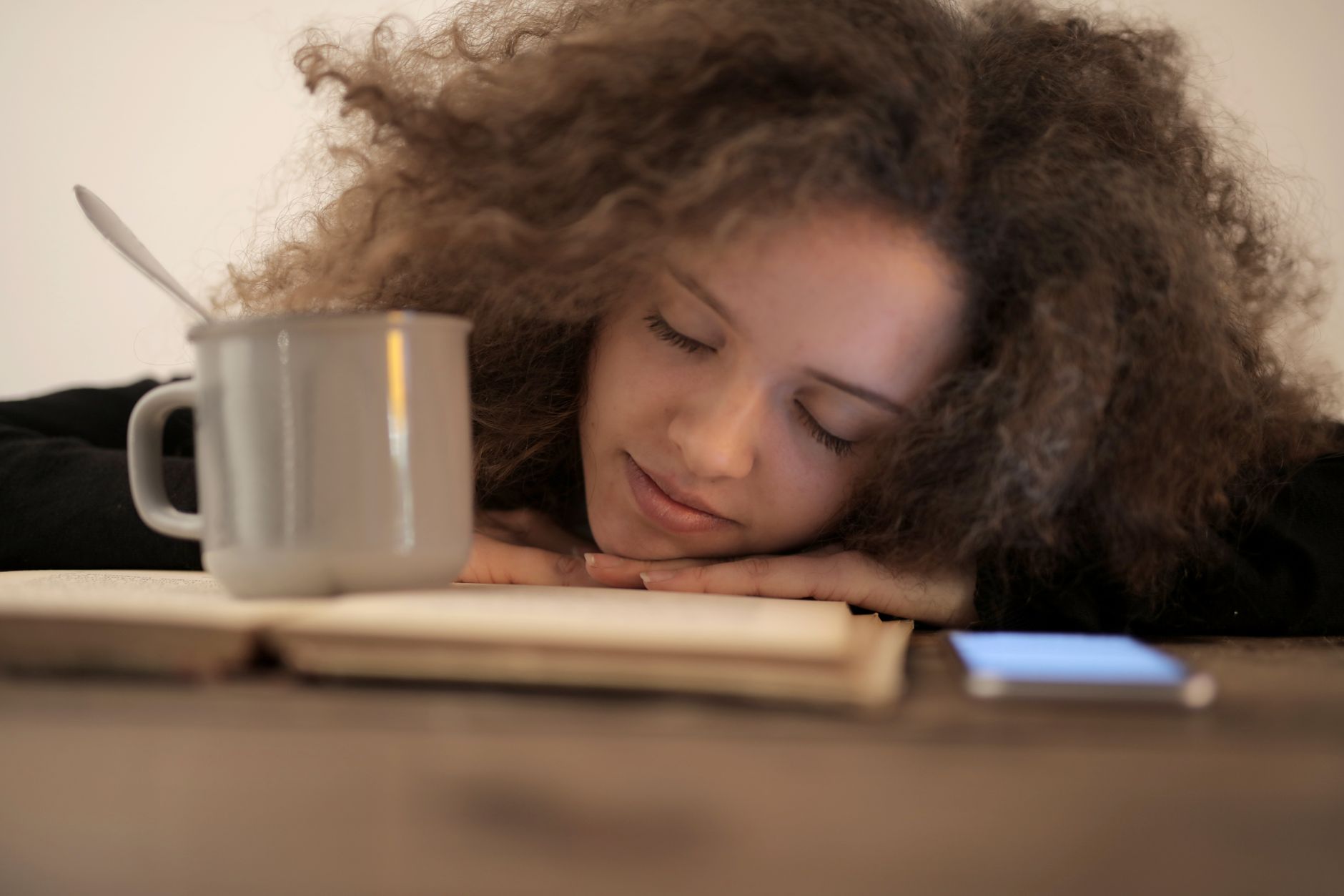 Como reduzir o tempo de tela no celular de verdade A young woman resting her head on a book with a mug nearby, relaxing indoors.