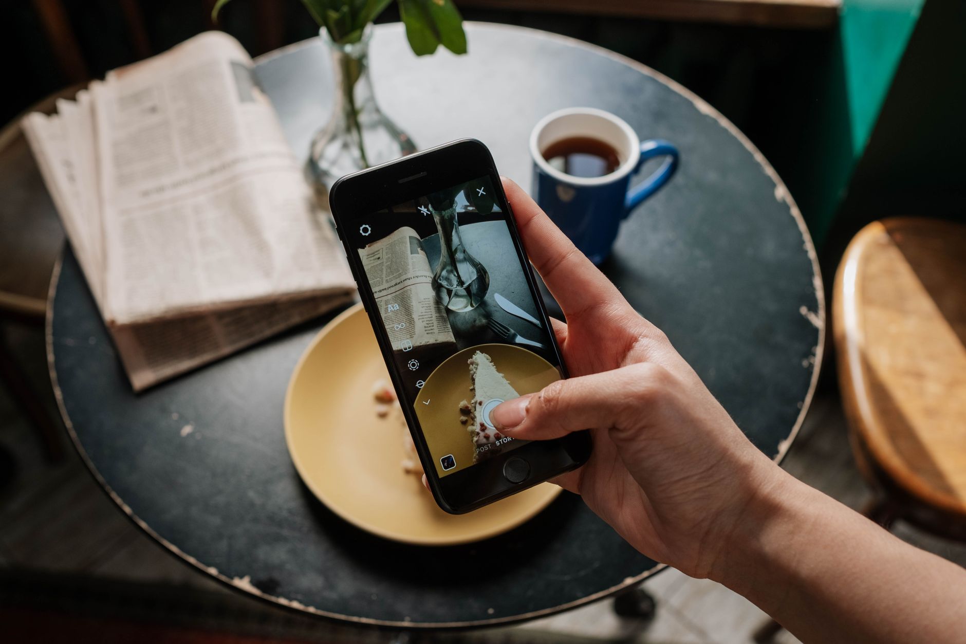 A person holding a smartphone to photograph a slice of cake in a warm café ambiance.