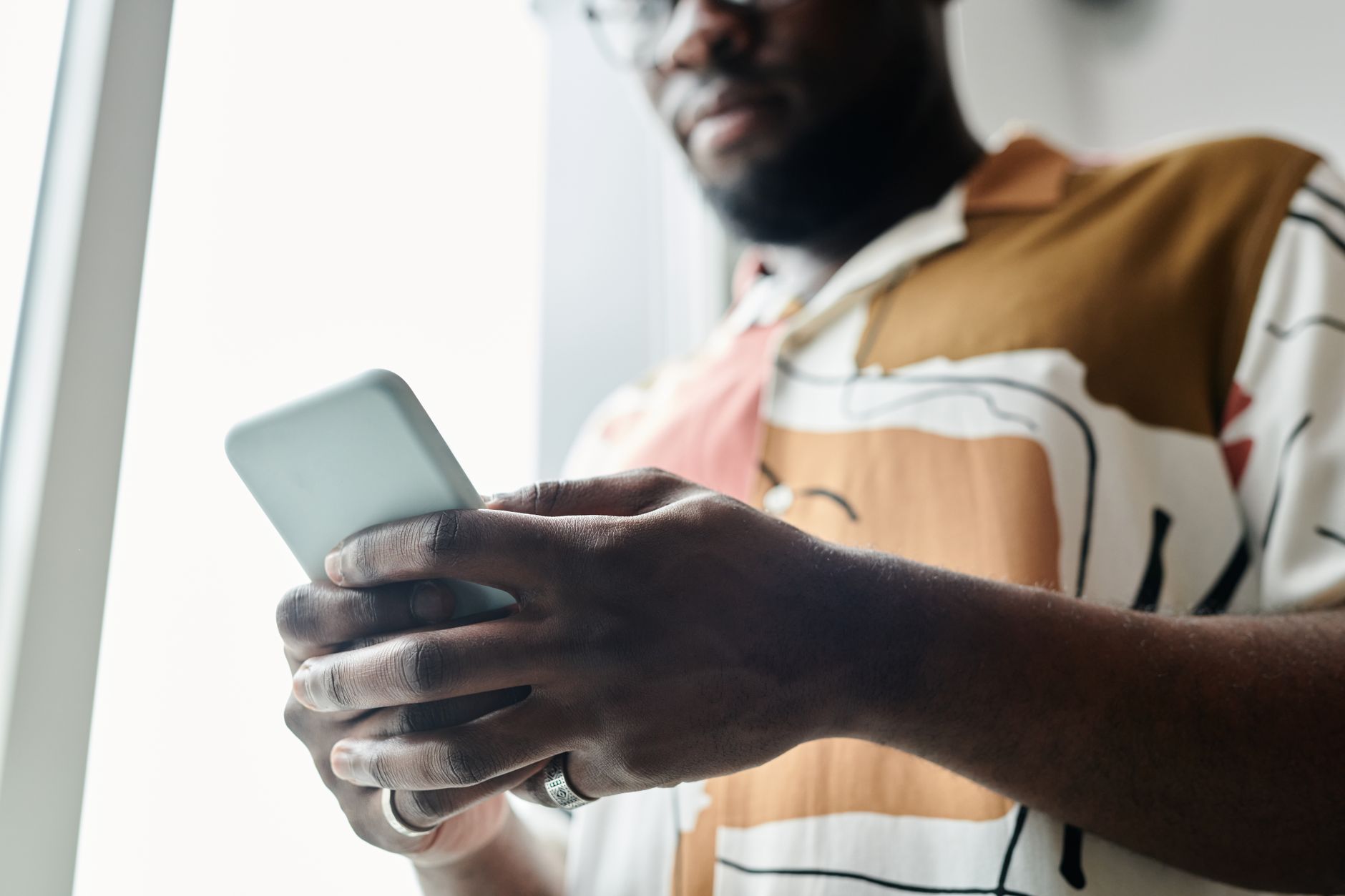 A close-up of a man using a smartphone by a window, featuring a modern patterned shirt.
