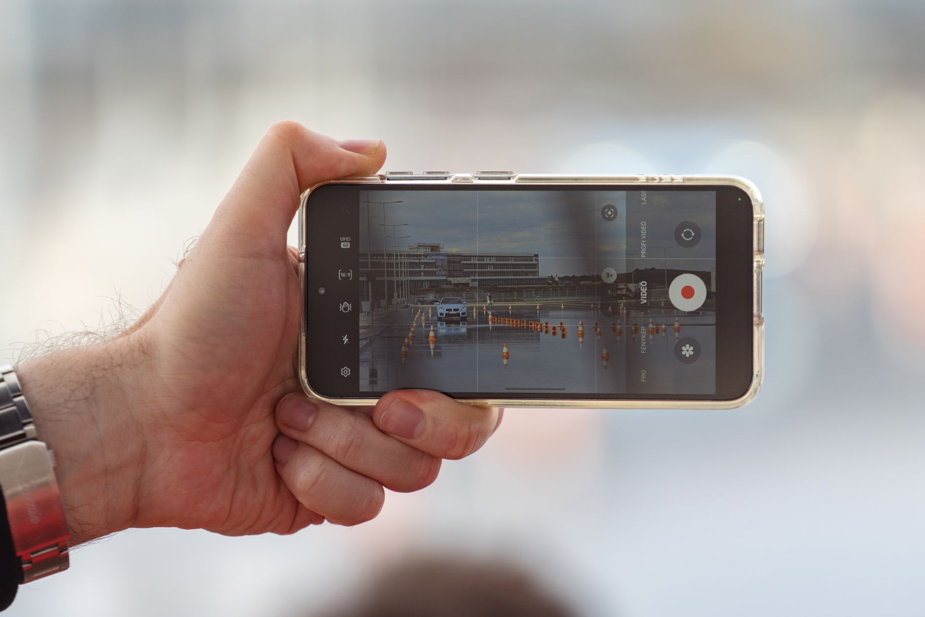 A hand holding a smartphone recording a video of a car scene outdoors. Captured in Balatonfőkajár, Hungary.