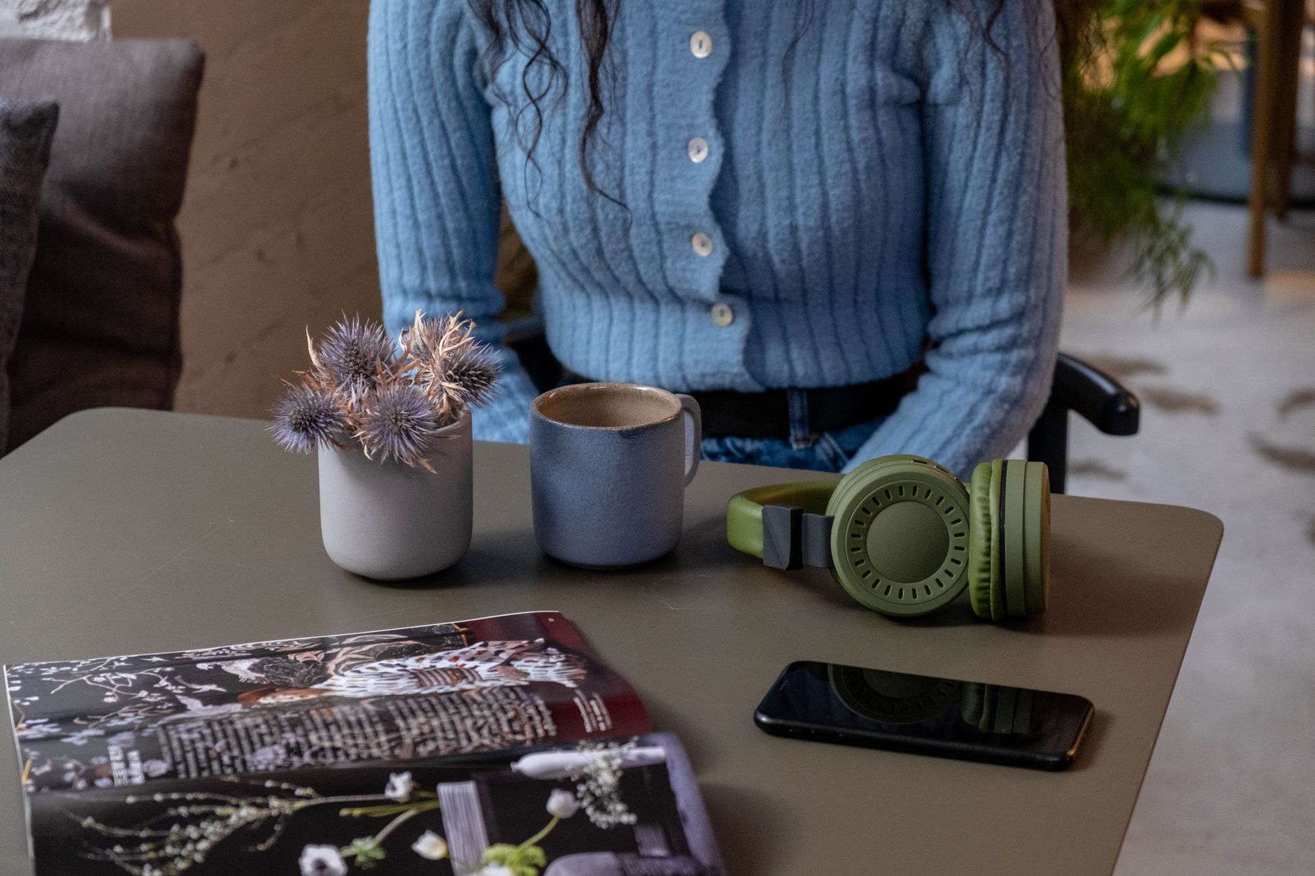 Como escolher entre fone Bluetooth ou com fio no celular A person in a blue sweater sits at a table with headphones, a phone, and a mug indoors.