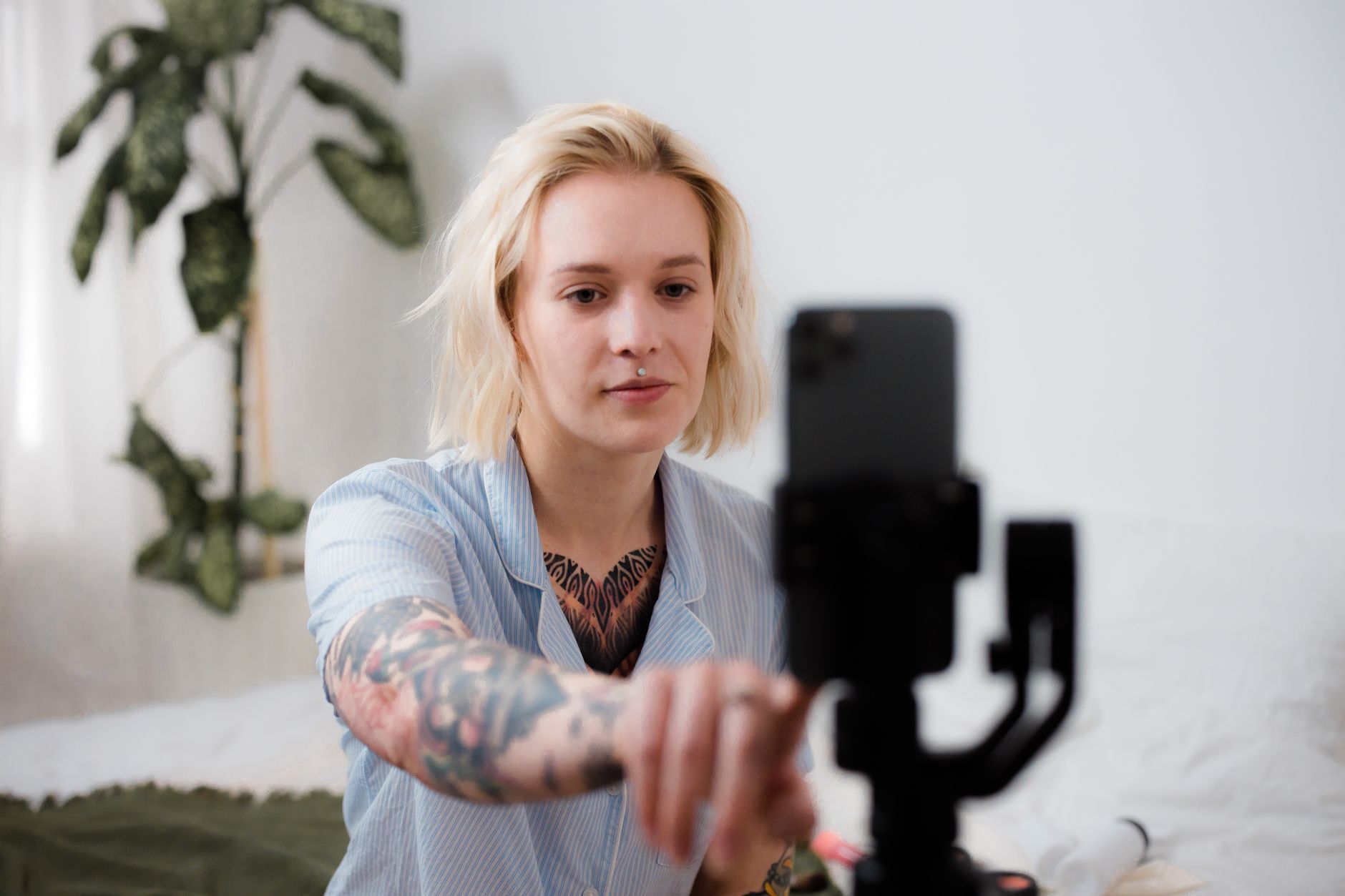 Galaxy S25 Ultra x iPhone 16 Pro Max: qual escolher? Tattooed woman with blonde hair adjusts smartphone on tripod for vlog setup indoors.