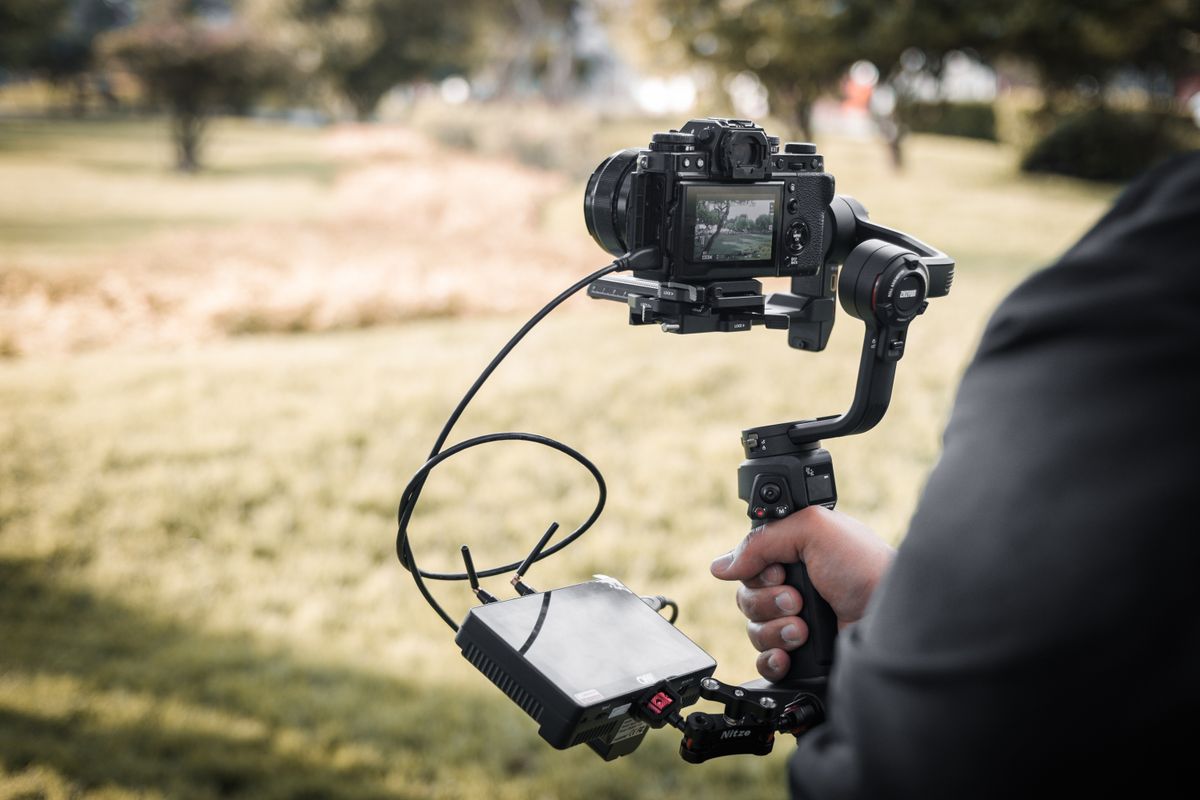 Pessoa segurando um gimbal mecânico com um smartphone acoplado gravando um vídeo em ambiente externo.