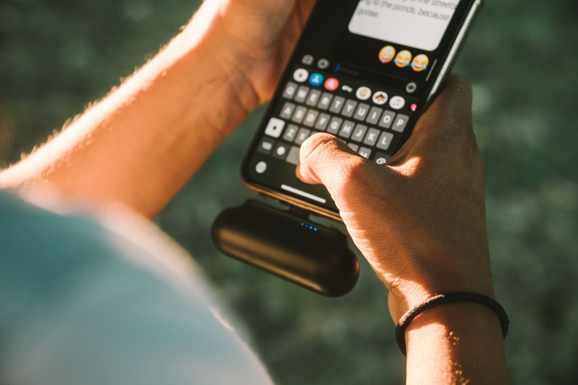 iPhone 12 ainda vale a pena em 2026? Close-up of a person texting on a smartphone connected to a power bank outdoors.