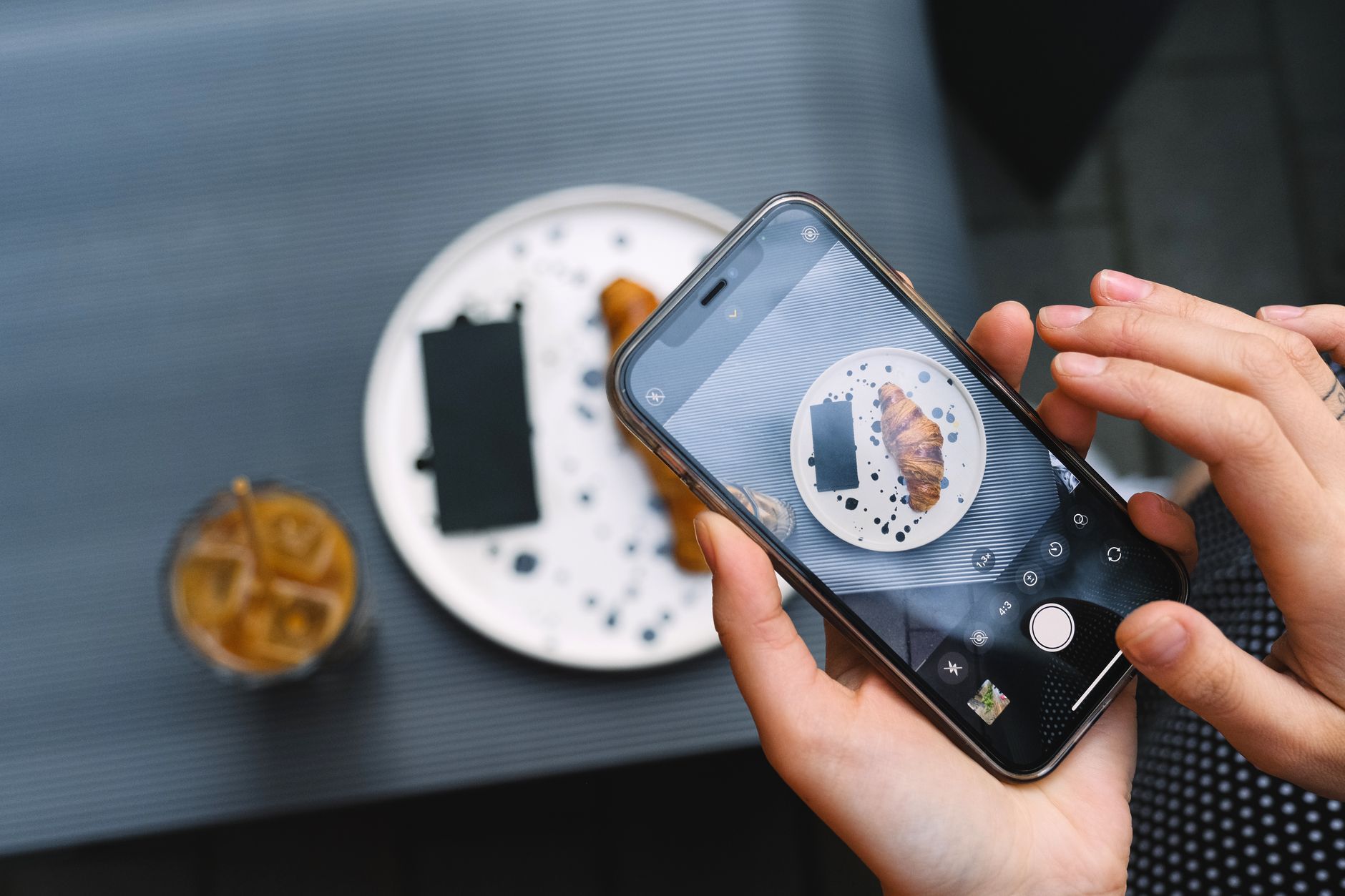 iPhone Pro x iPhone comum: diferenças que importam Hands photographing a croissant on a plate with a smartphone, featuring coffee.