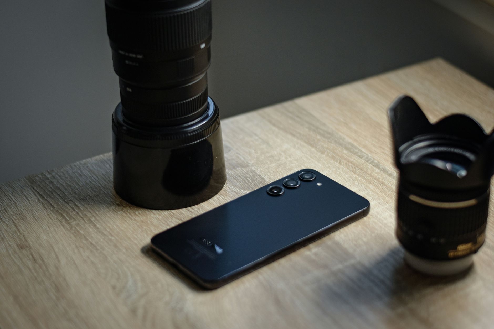 A sleek smartphone placed alongside camera lenses on a wooden desk, capturing technology and photography.