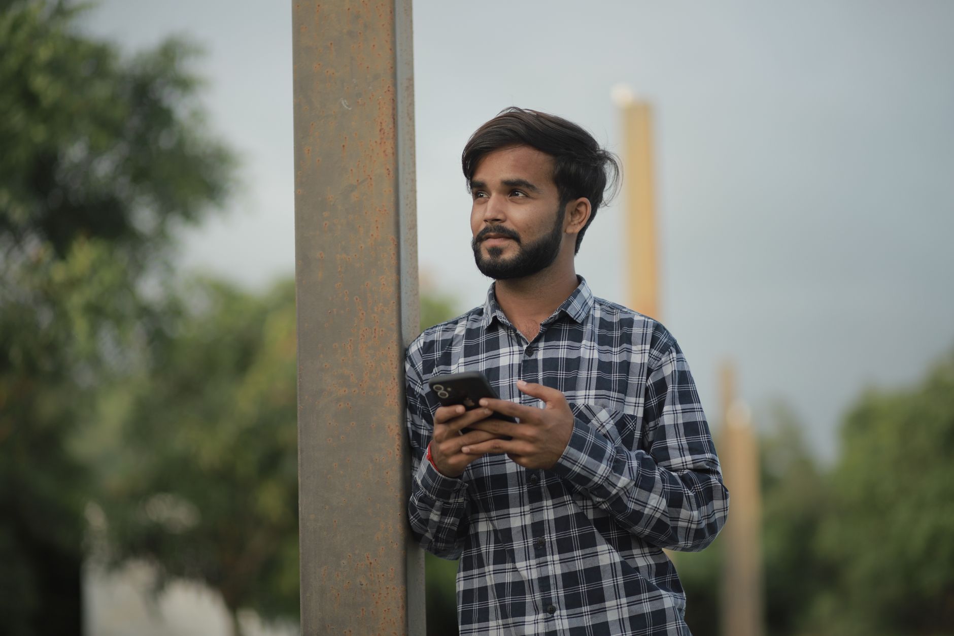 Melhor celular até mil reais: como escolher A young bearded man in a checked shirt checks his smartphone outdoors in a casual portrait.