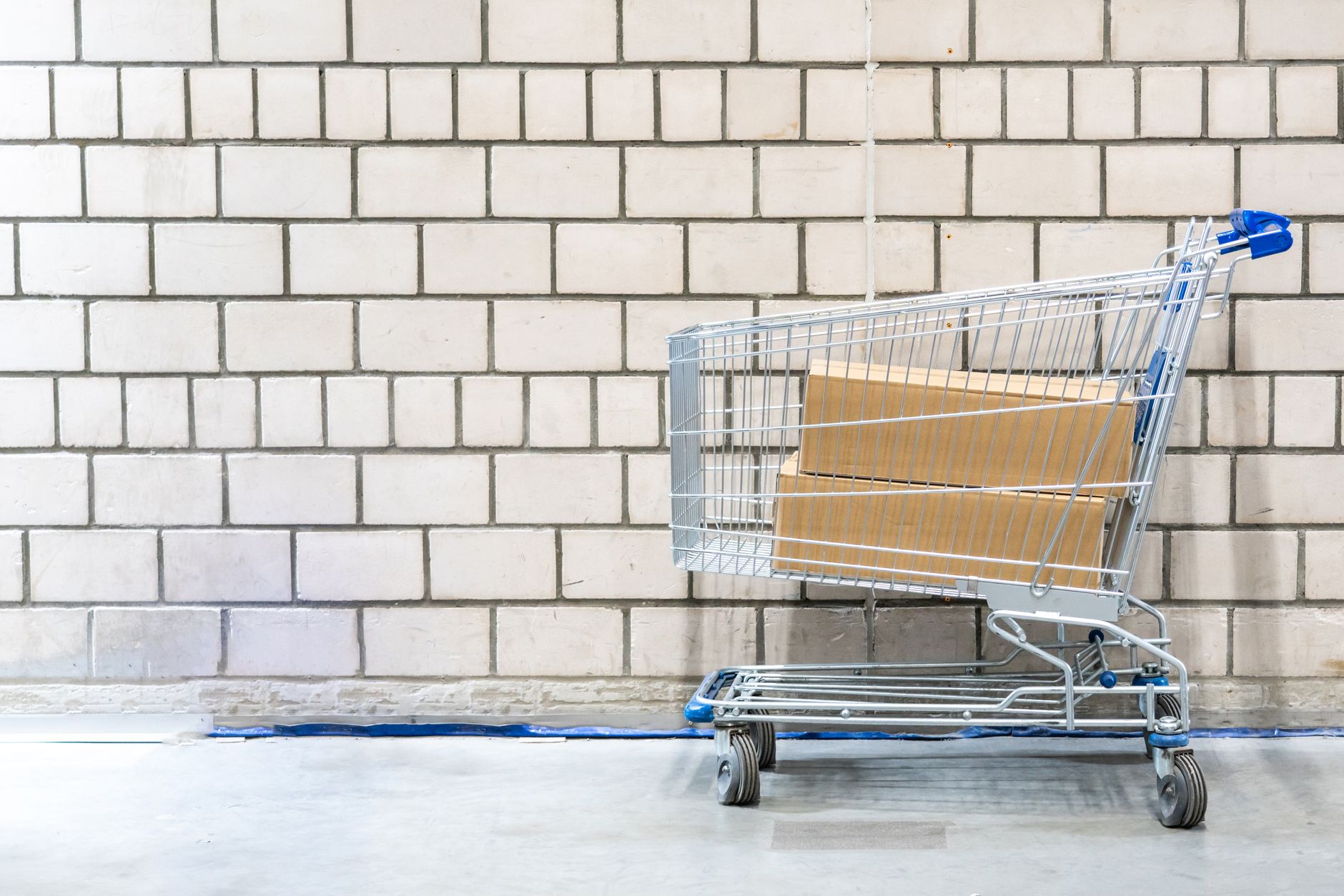 Por que o celular é tão caro no Brasil? Shopping cart with cardboard boxes against a concrete block wall. Minimalist and industrial setting.