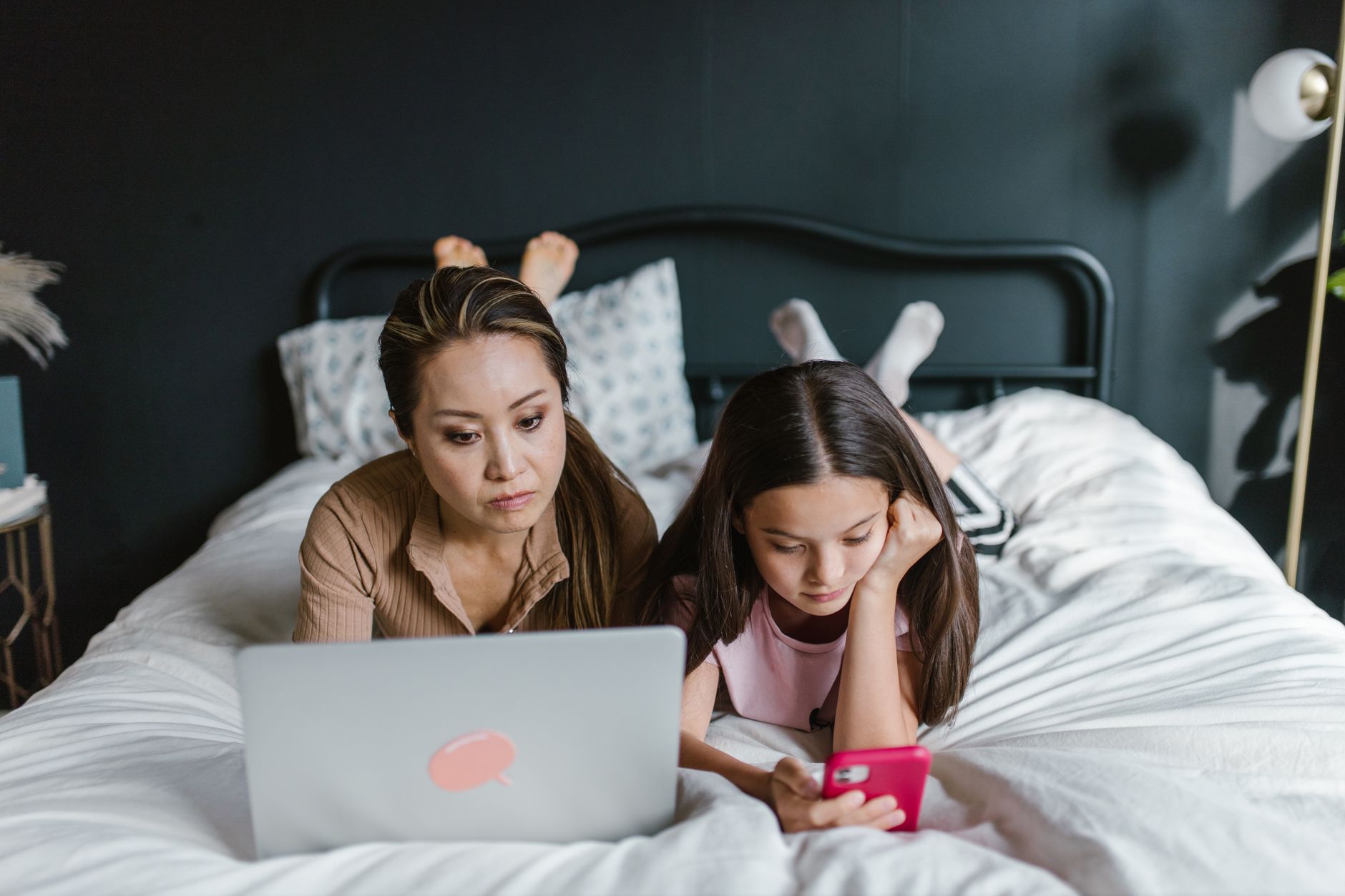 Mother and daughter lying on bed using laptop and smartphone indoors, depicting modern family life.