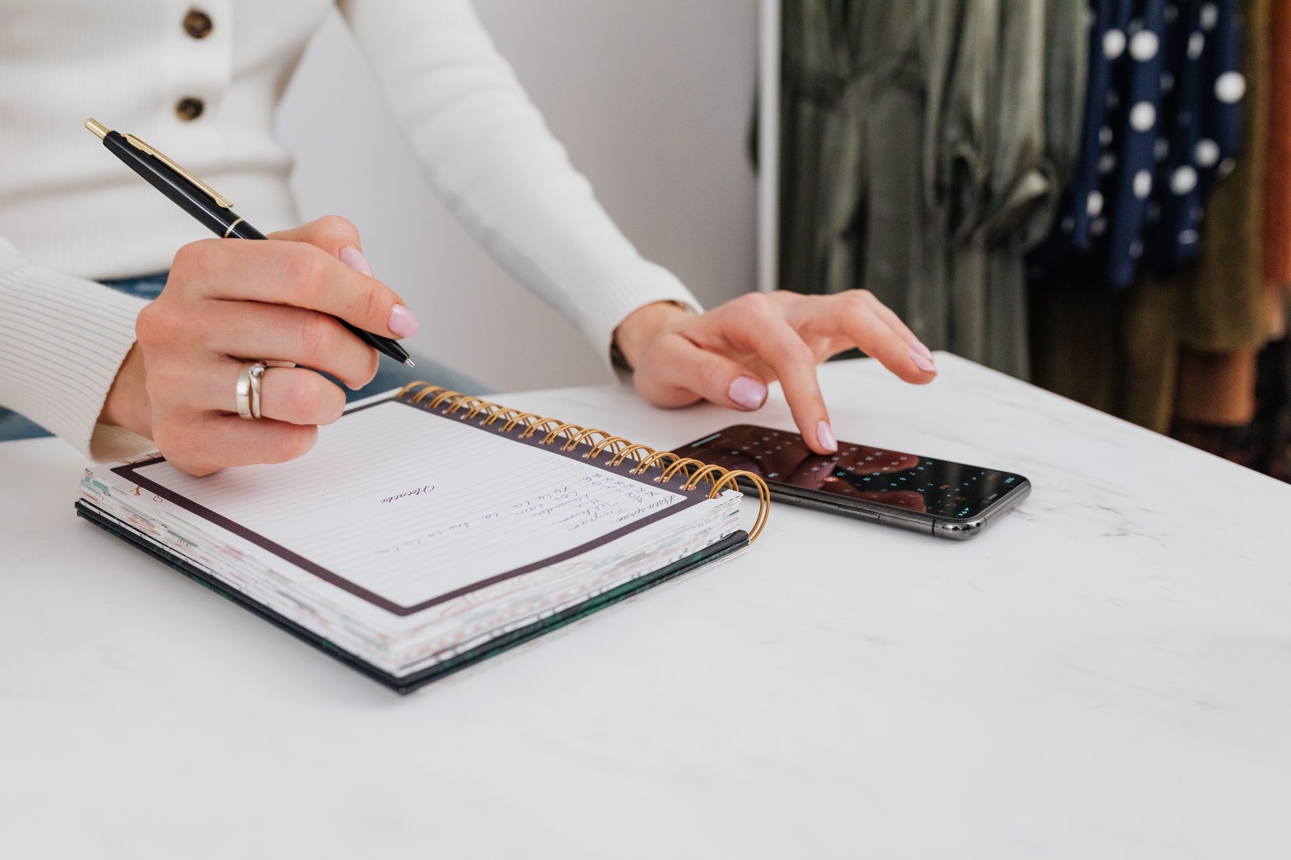 Qual é a melhor época para comprar celular? Close-up of hands writing in a notebook, with a smartphone on the table.