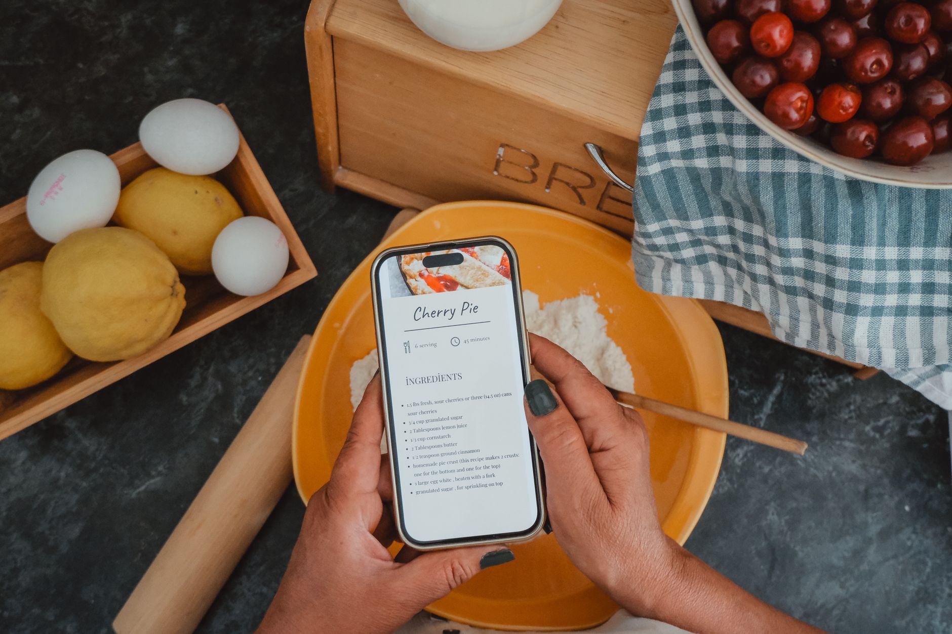 Quanto tempo esperar para o celular baixar de preço Top view of hands holding a phone with a cherry pie recipe, surrounded by baking ingredients.