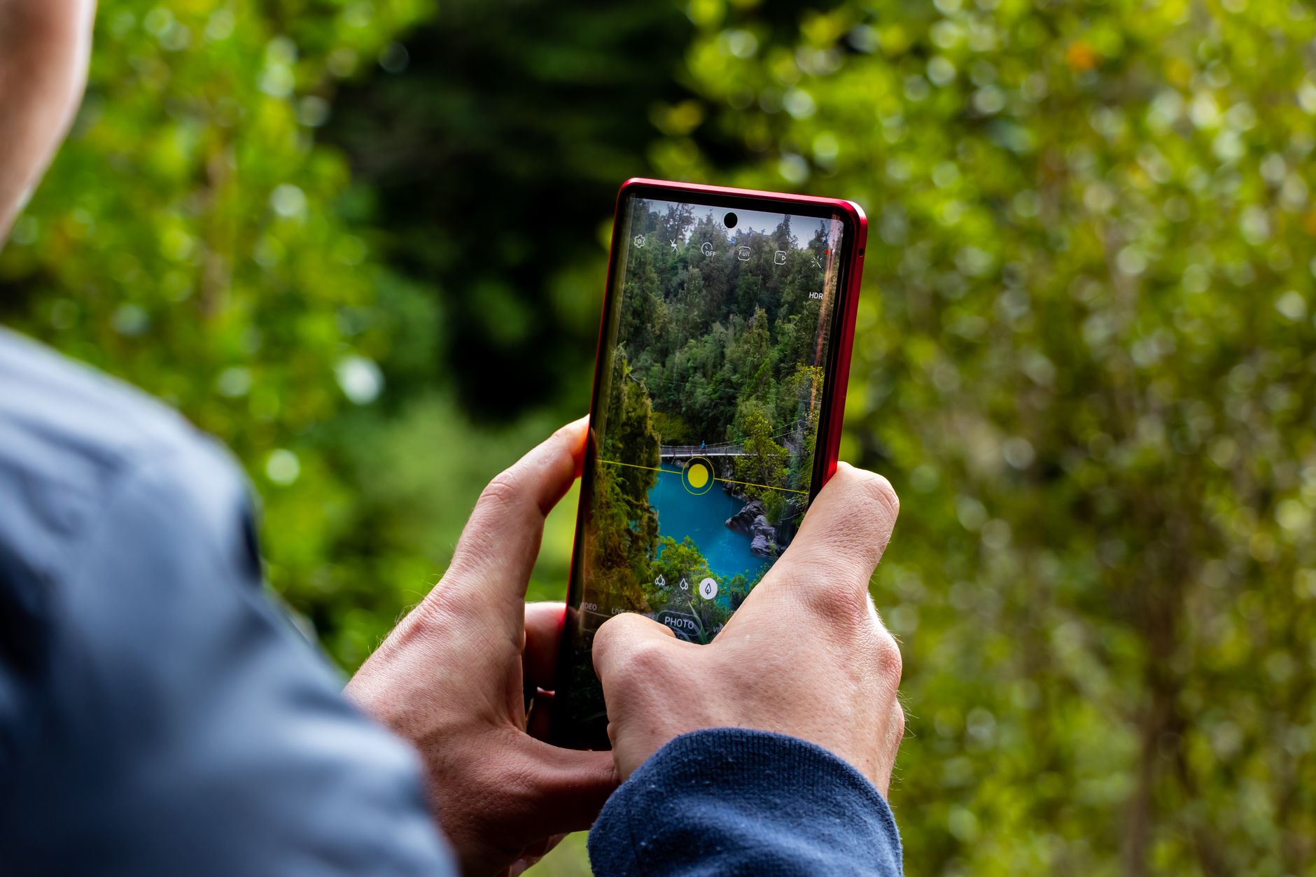 Quanto vale seu celular usado: como calcular Hands holding a smartphone capturing a scenic river view with trees.