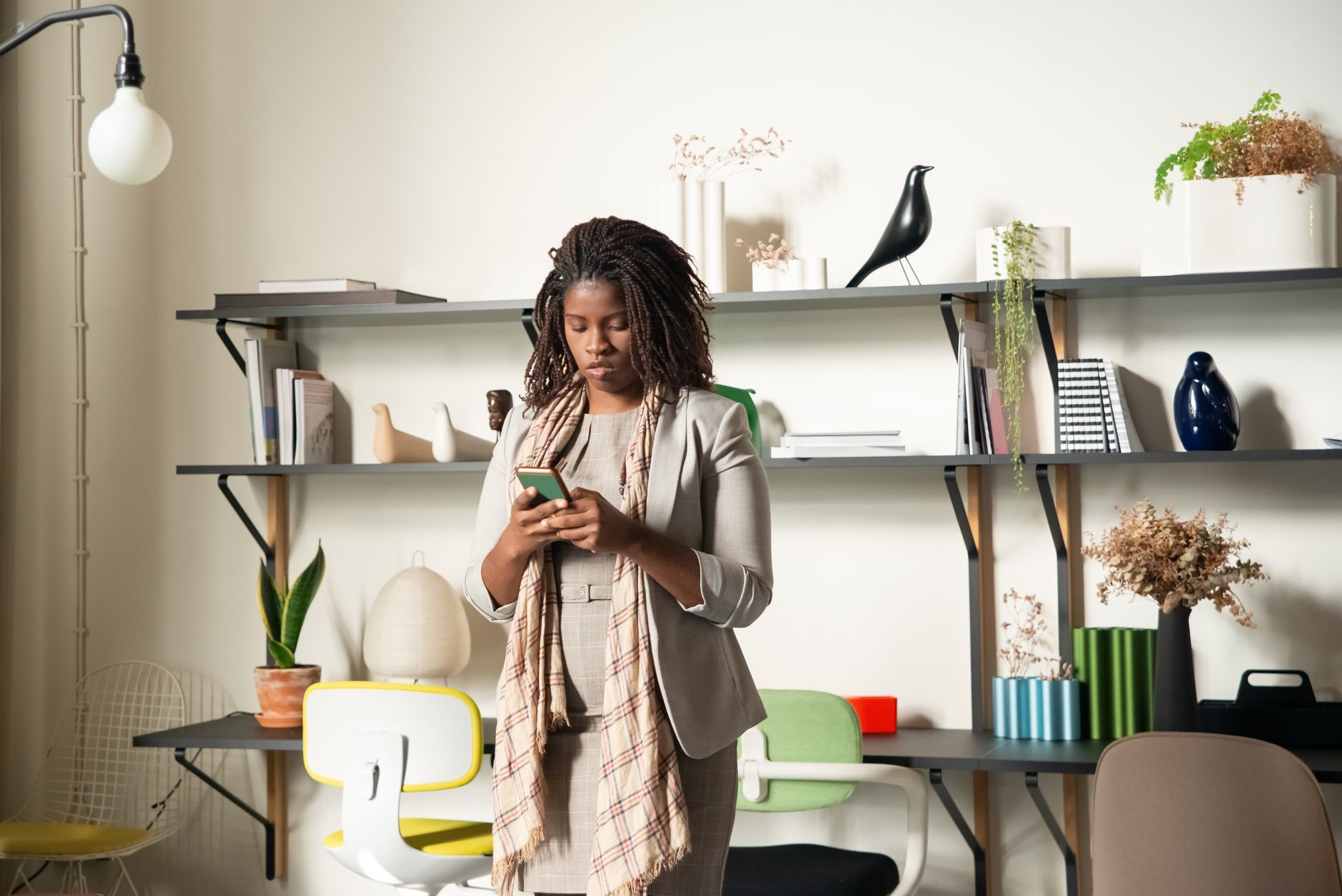 128GB, 256GB ou 512GB: quanto armazenamento escolher A woman standing in a modern office environment, focused on her smartphone, surrounded by books and plants.