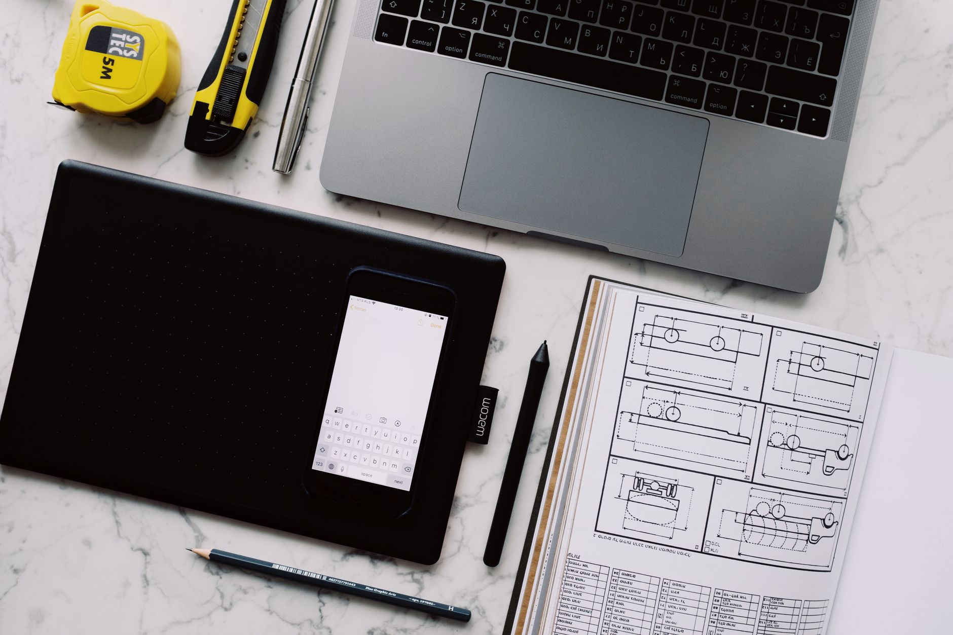 Como escolher entre tablet ou celular de tela grande From above view of modern workplace at white marble table with various gadgets and tools and opened notebook with drawings with pen and pencil