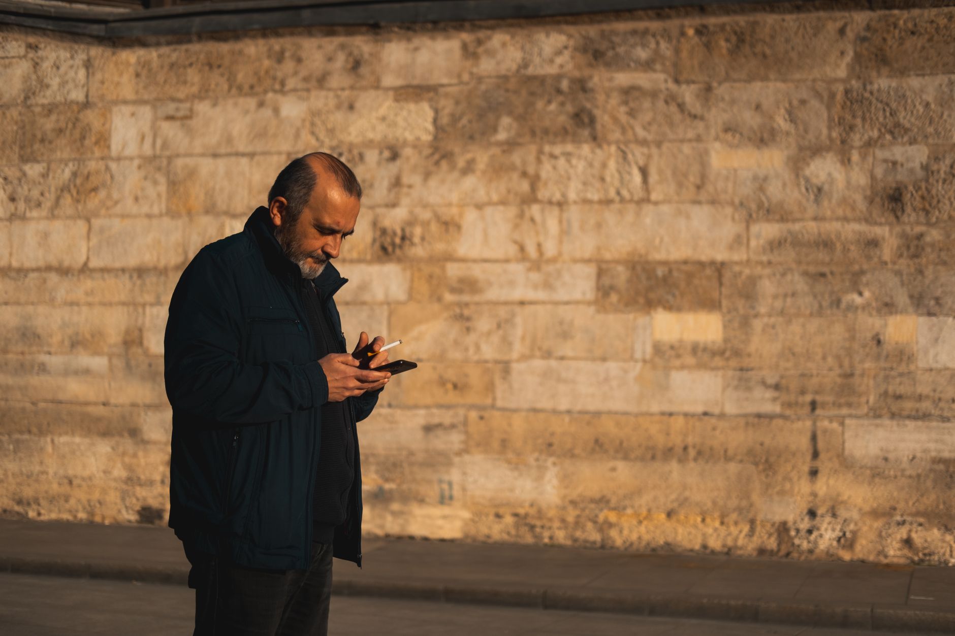 Quanto valem cem euros em reais? Veja como calcular Man standing on sidewalk using smartphone near a rustic stone wall.