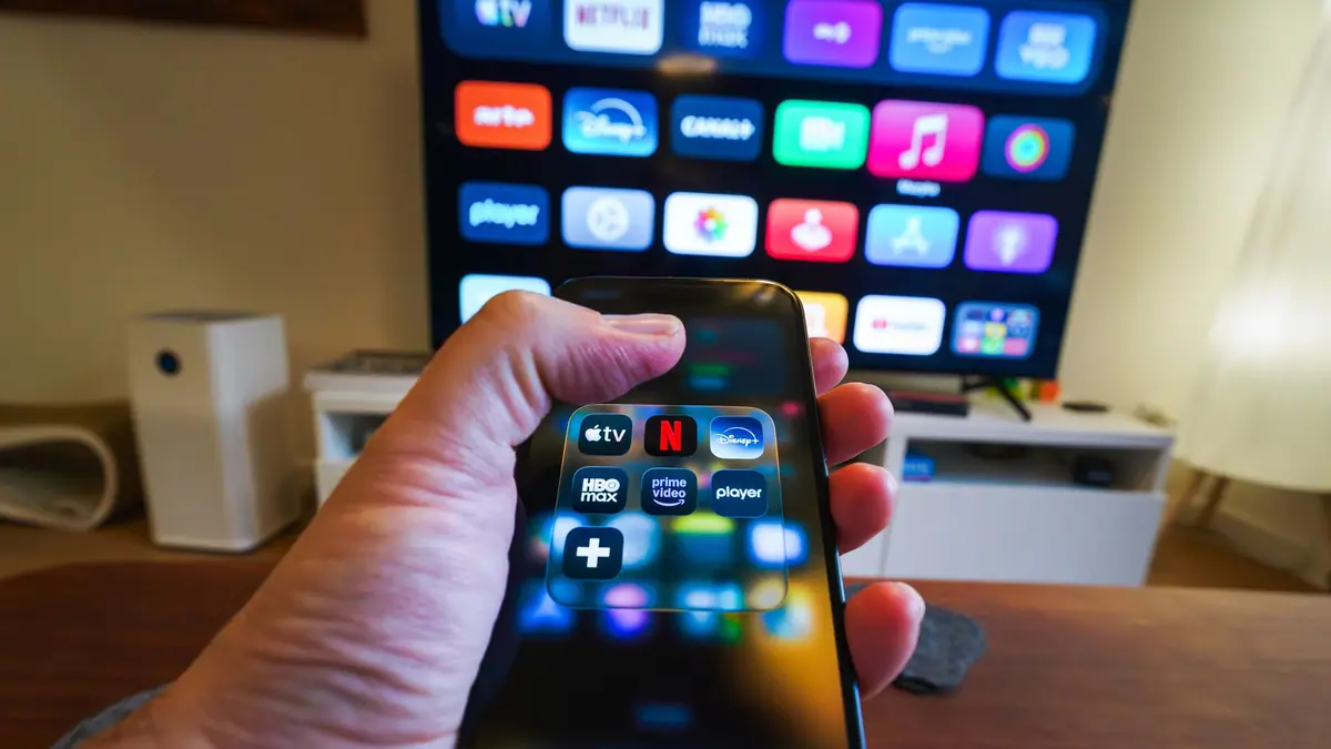 Mão segurando um celular com aplicativos de streaming diante de uma TV
