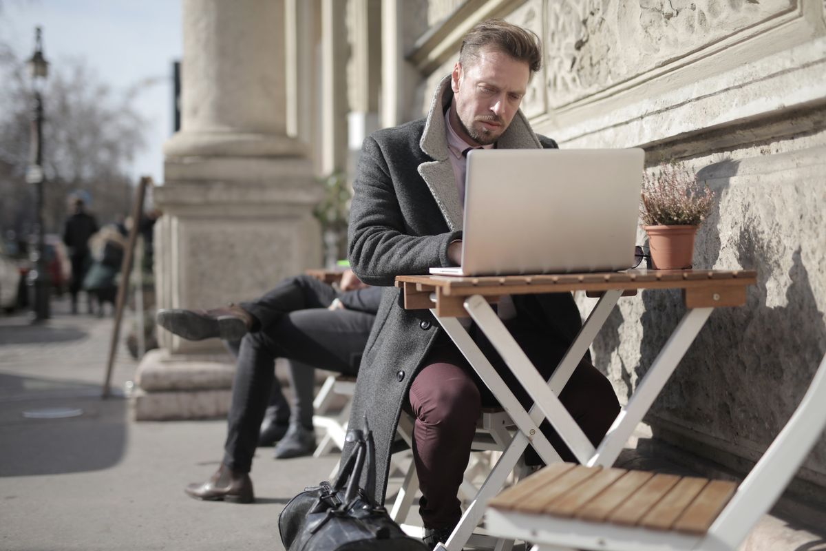 Pessoa segurando cartão de débito e celular em uma mesa de café na Europa com notebook ao lado.