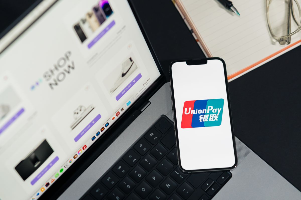 Pessoa segurando celular com aplicativo bancário aberto exibindo dados de cartão virtual ao lado de um notebook em mesa de trabalho.