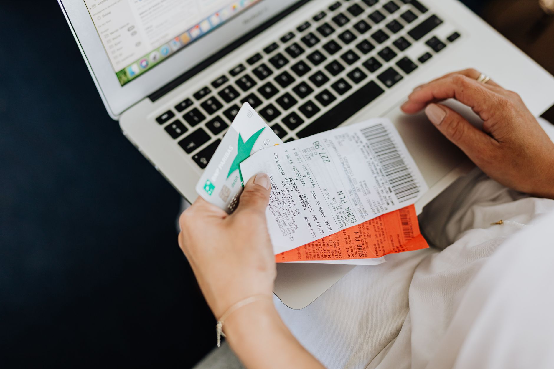 Como comparar taxas entre cartões internacionais Close-up of hands holding receipts and a bank card in front of a laptop, representing online shopping and e-commerce.