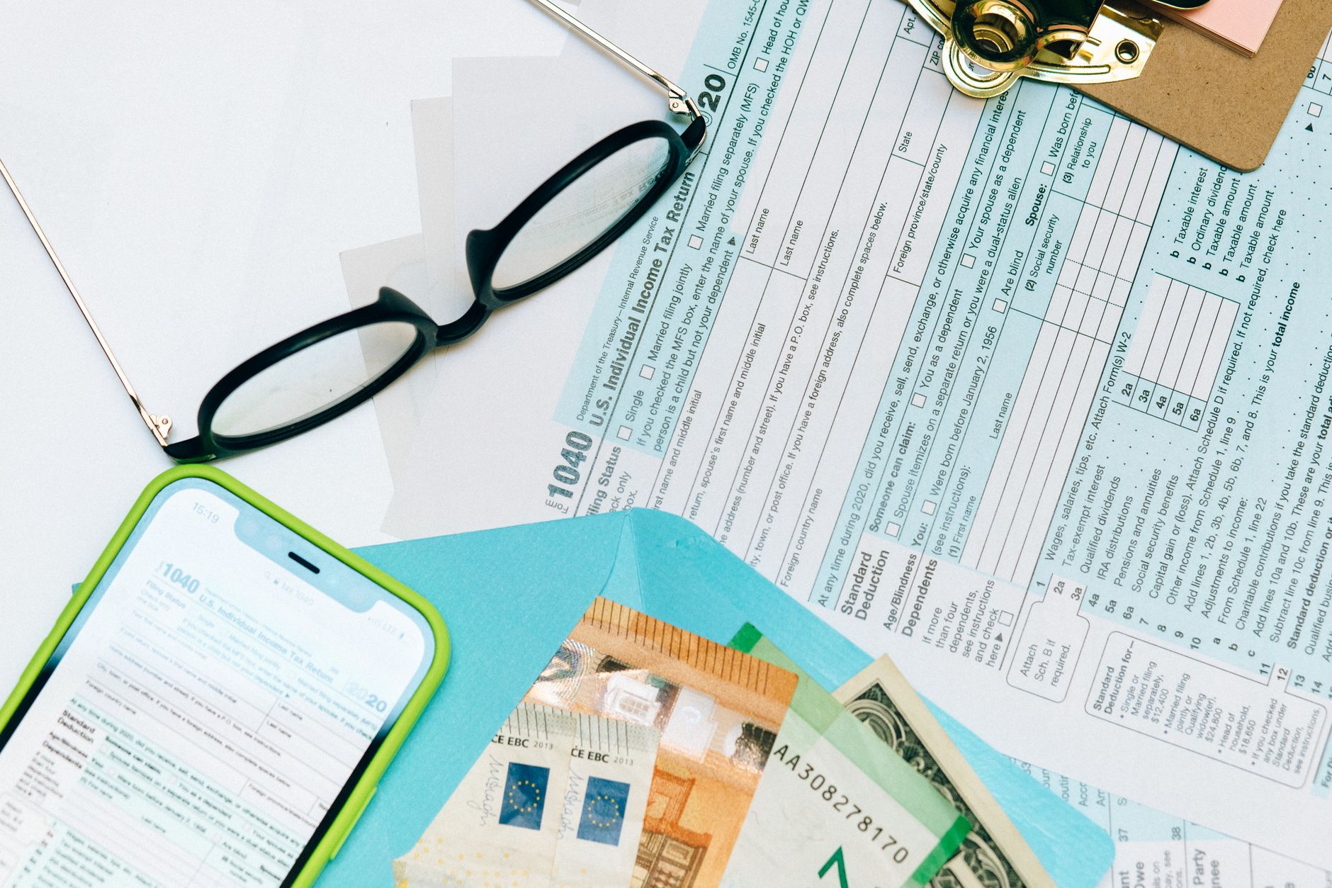 Pessoa preenchendo declaração de imposto de renda no computador com passaporte e cédulas de dólar sobre a mesa.