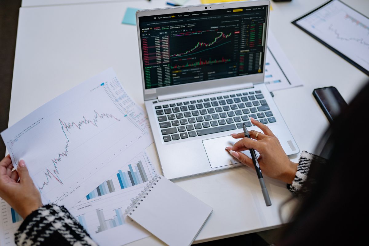 Pessoa analisando gráficos financeiros e notebook com notas de euro sobre a mesa.