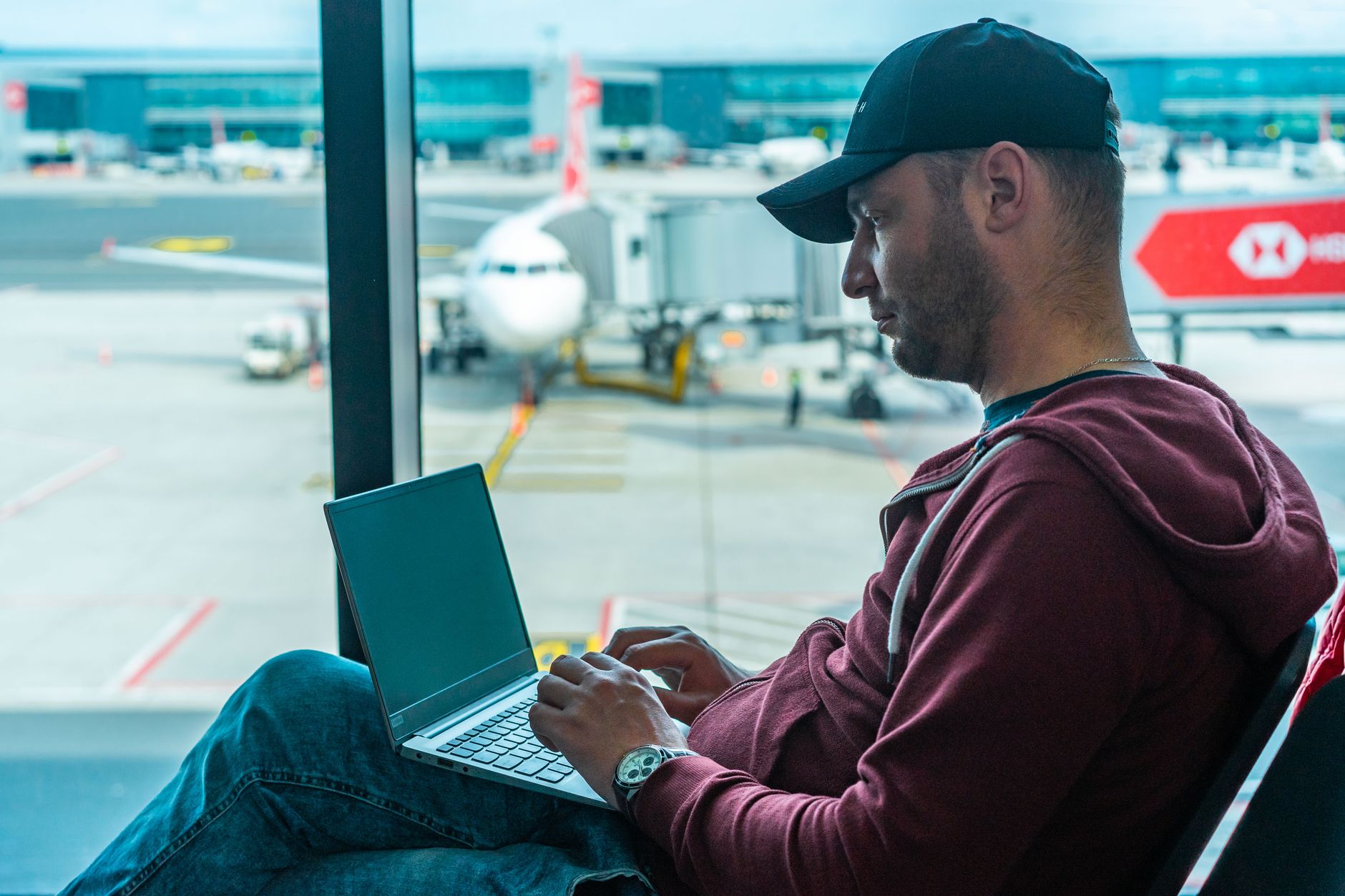 Como comprar dólar antecipado para viajar A man working on his laptop in an airport terminal, with a plane visible outside.