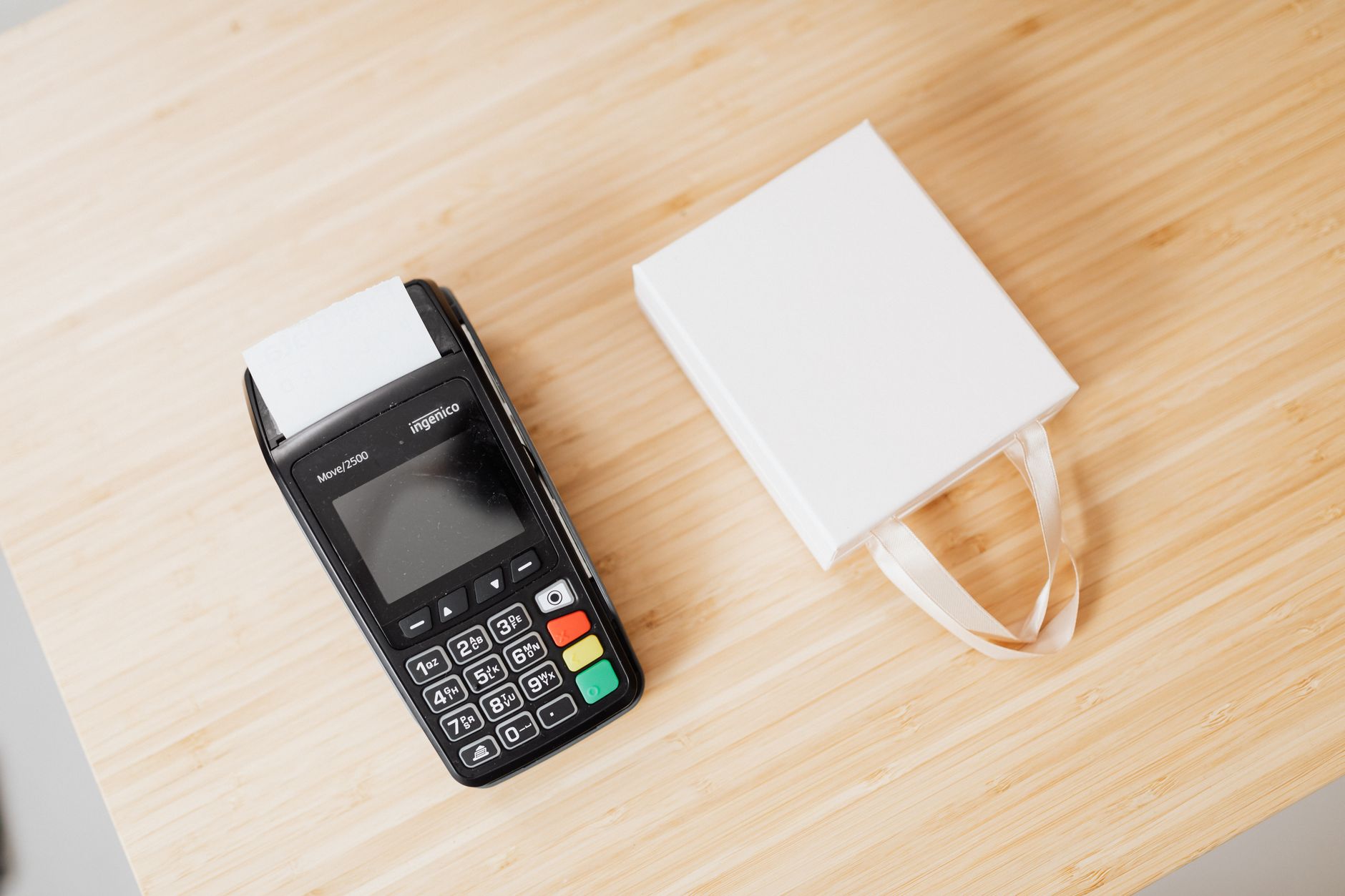 Como funciona o extrato de uma conta em euro A payment terminal and a small white gift bag on a wooden countertop, ideal for commerce themes.