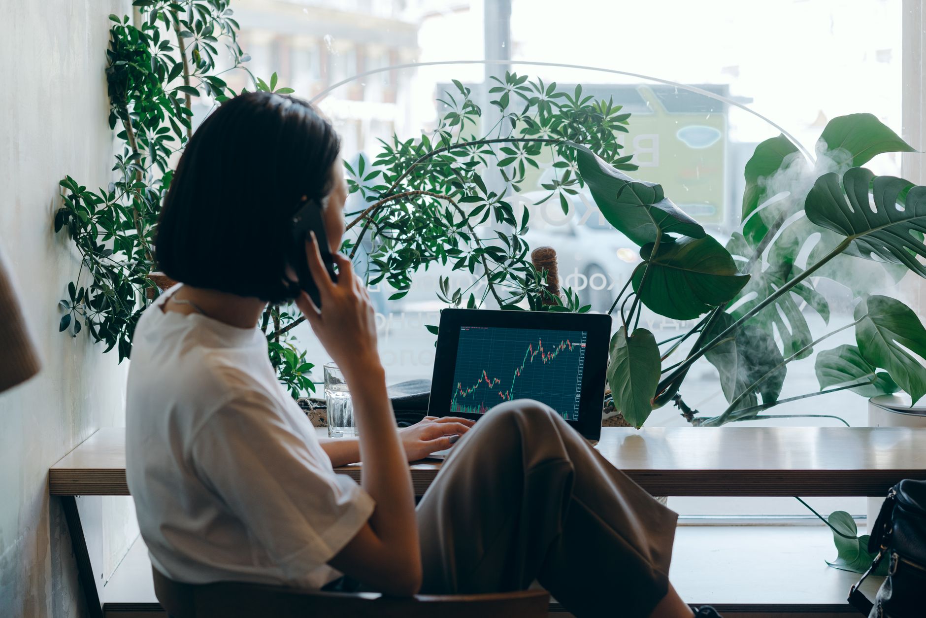 Businesswoman on phone, analyzing stock graphs on laptop in a café.