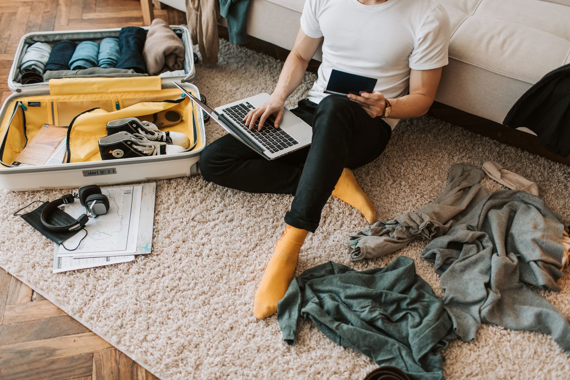 Man sits on floor packing suitcase while using a laptop, balancing travel and remote work.