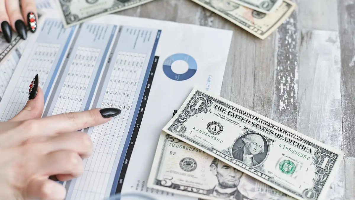 Mãos analisando planilha financeira com notas de dólar sobre a mesa