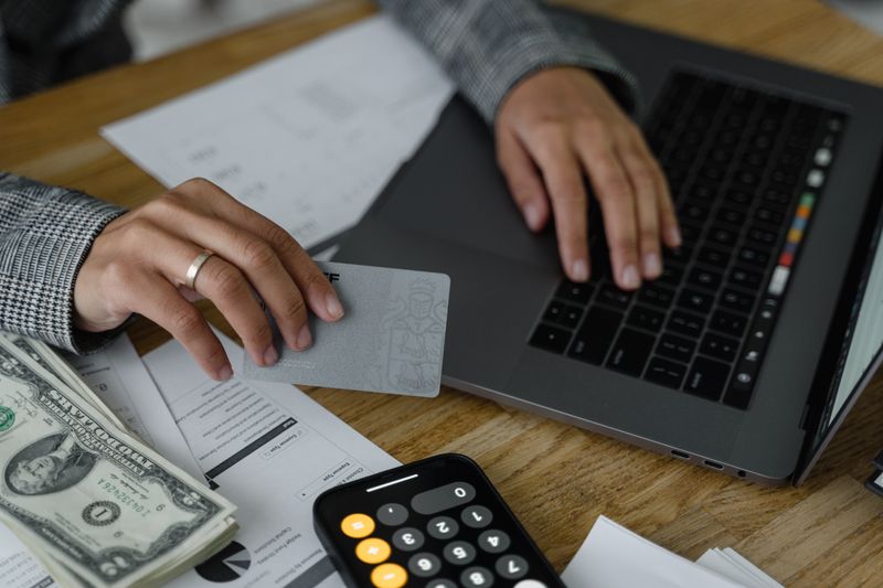 Dólar para Viagem: Comprar de Uma Vez ou Parcelado? Close-up of a person using a credit card and laptop in a financial setting with cash and calculator.