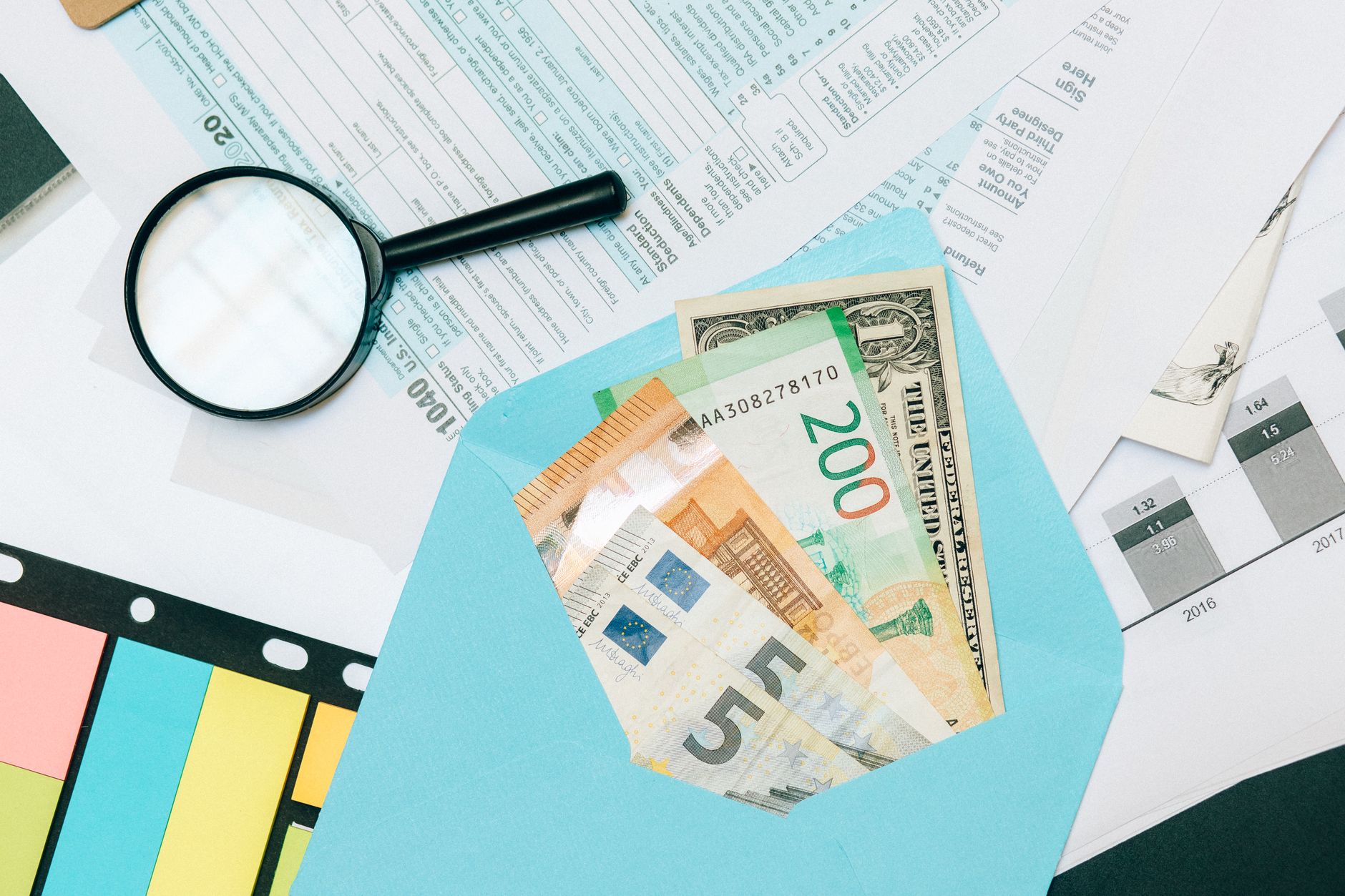Qual foi a menor cotação do euro frente ao dólar? Close-up of tax documents and mixed currency in a blue envelope with a magnifying glass, perfect for financial analysis concepts.