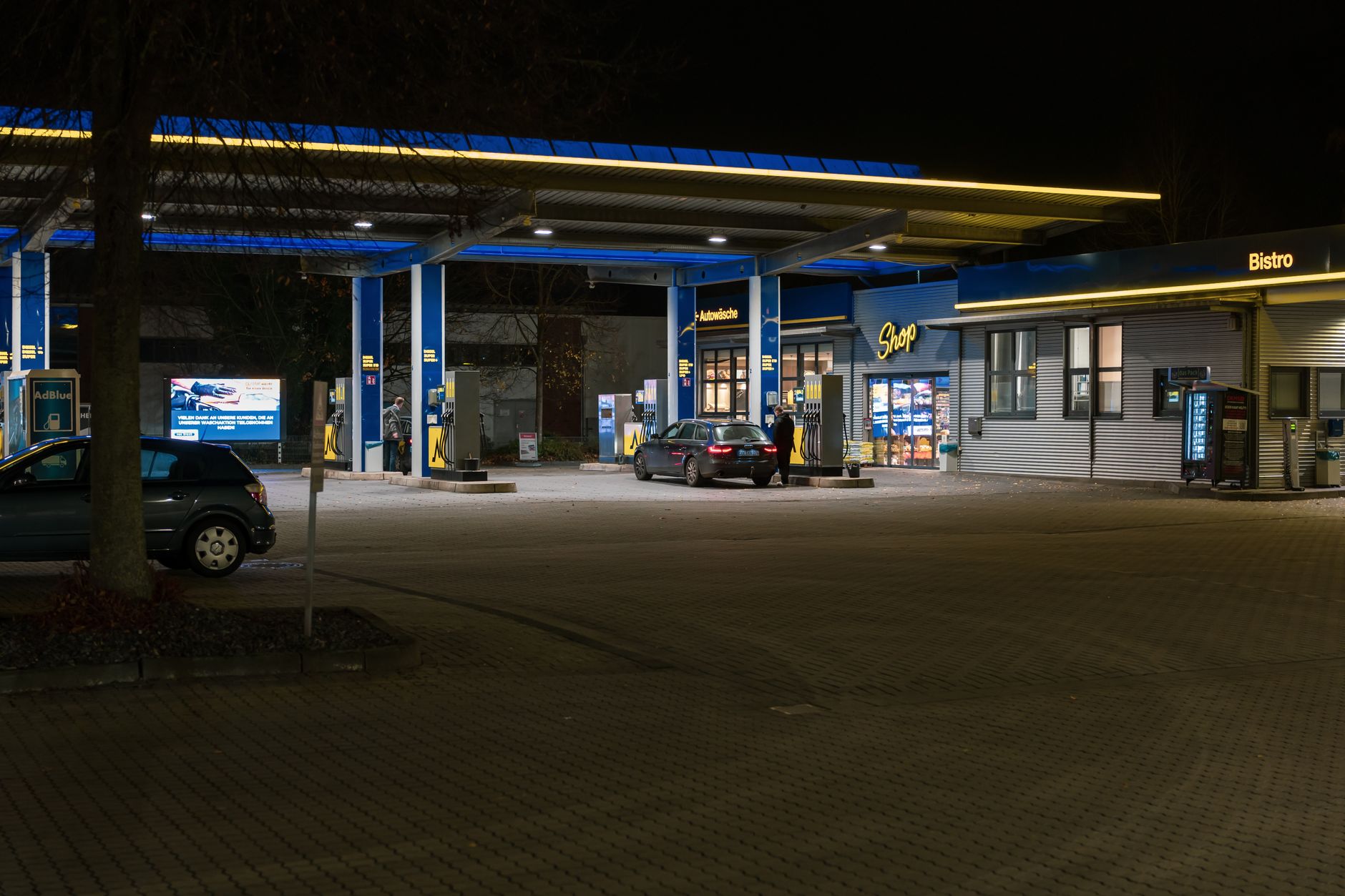 A well-lit gas station at night featuring cars and a bistro, creating a lively urban scene.