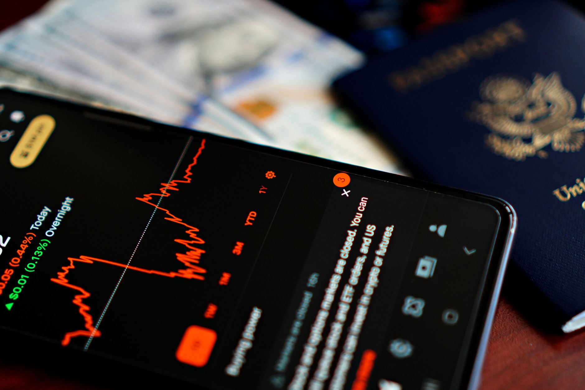 Moedas de euro e notas sobre uma mesa ao lado de um passaporte e um smartphone mostrando gráficos econômicos.