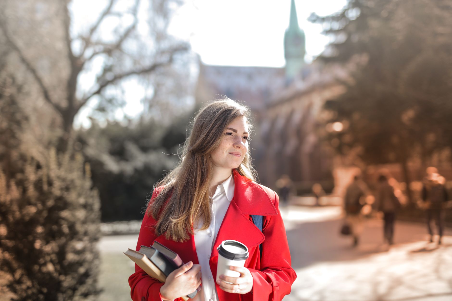 Euro na Carteira para Custear Estudos de Filhos na Europa Smiling woman in red coat with coffee and books, enjoying leisure time outdoors on a university campus.