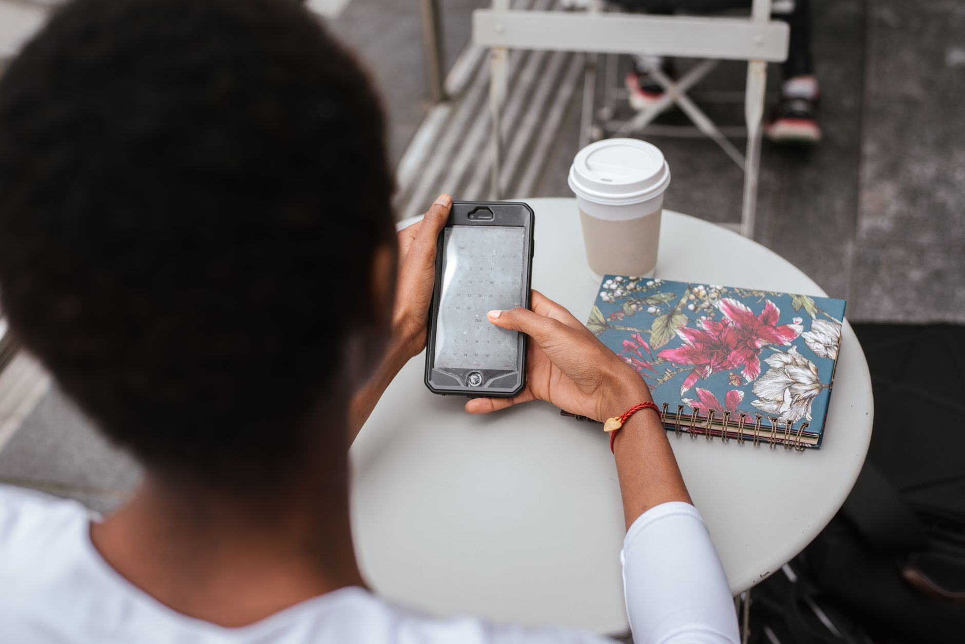 High angle back view of faceless African American lady sitting at table in cafeteria and checking notification on smartphone near notebook and cup of drink