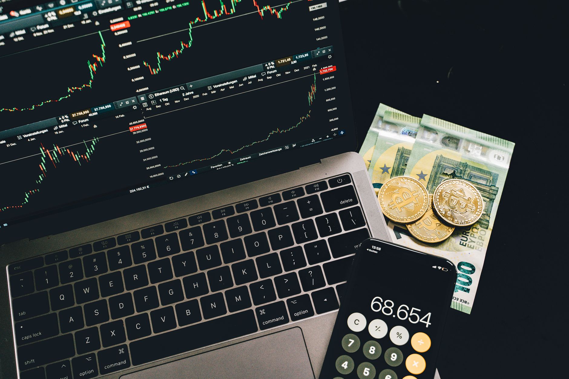 Euro-real histórico: como entender a cotação ao longo do tempo A laptop displaying stock charts with Bitcoin, Euros, and a cellphone calculator, showcasing financial analysis.