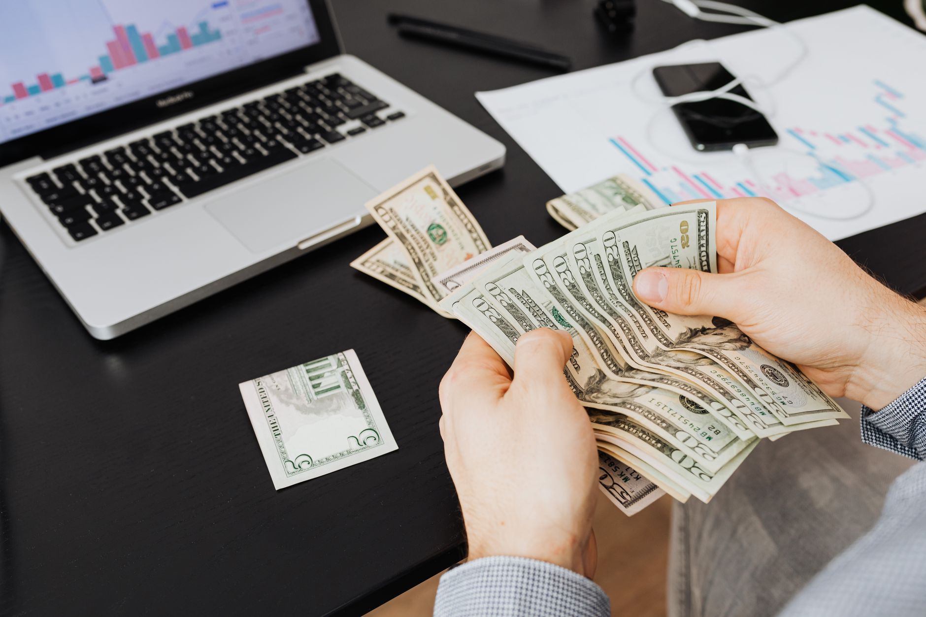 Remessa internacional em euro paga imposto? Person counting dollar bills at desk with laptop and financial charts, symbolizing finance management.