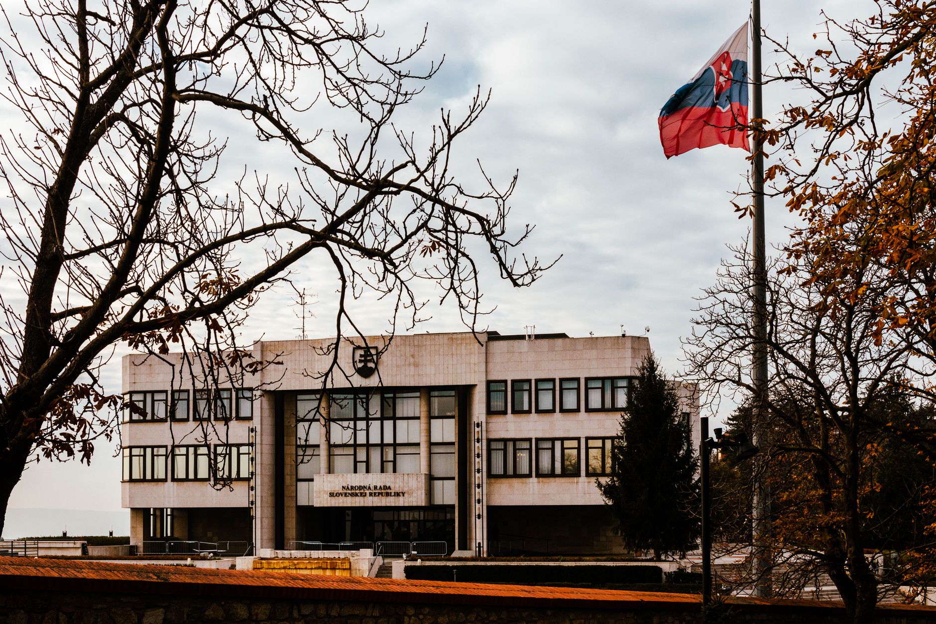 Fed x BCE: como os bancos centrais afetam o euro A view of the Slovak Parliament building surrounded by autumn trees with the national flag waving under an overcast sky.