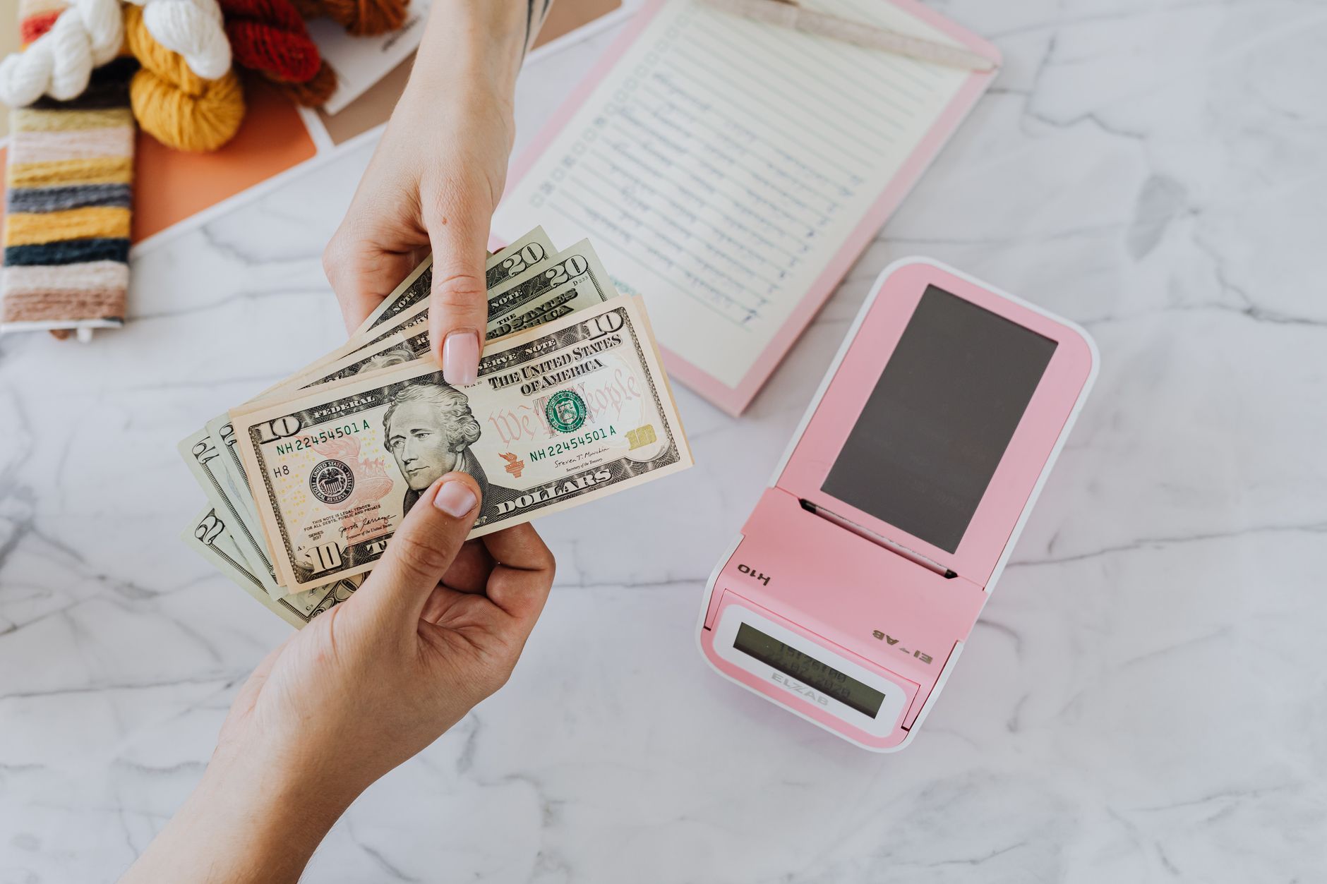 É melhor guardar dinheiro em euro ou dólar? Close-up of hands exchanging US dollars with a pink calculator on a marble surface.