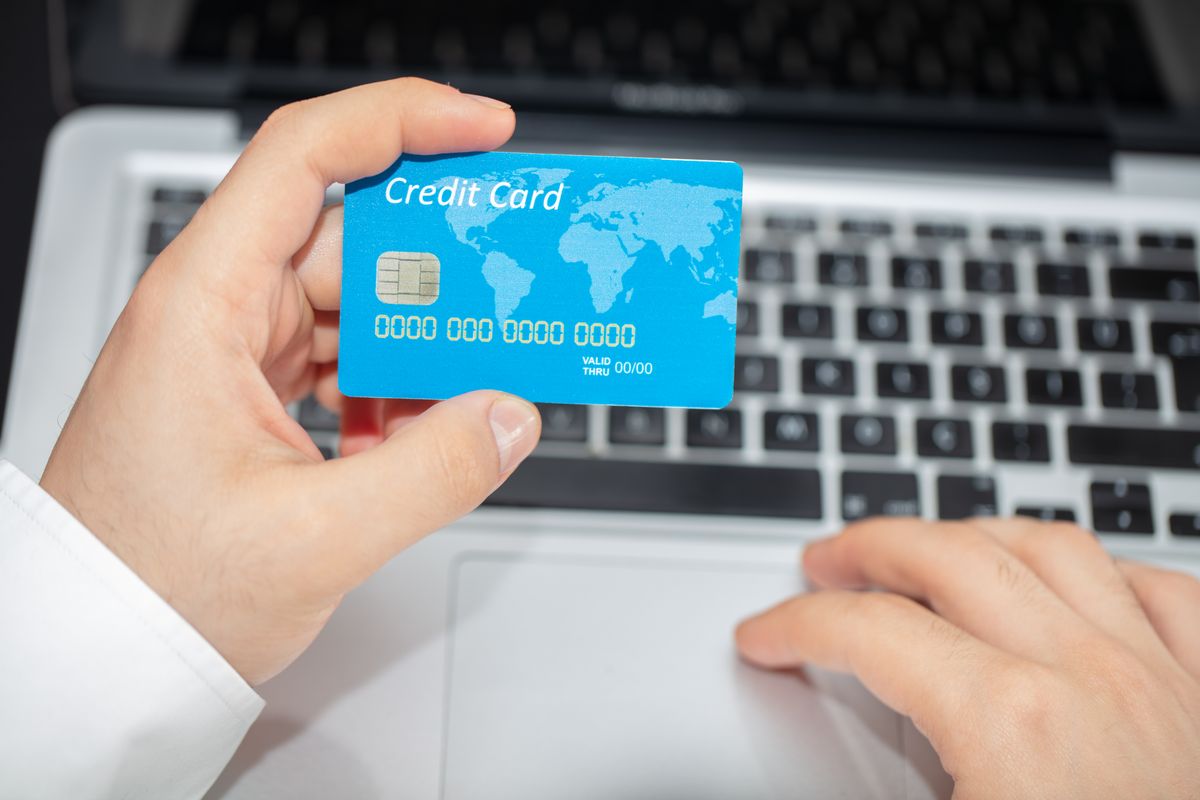 Pessoa segurando cartão de crédito internacional em frente a um computador navegando em site de finanças.