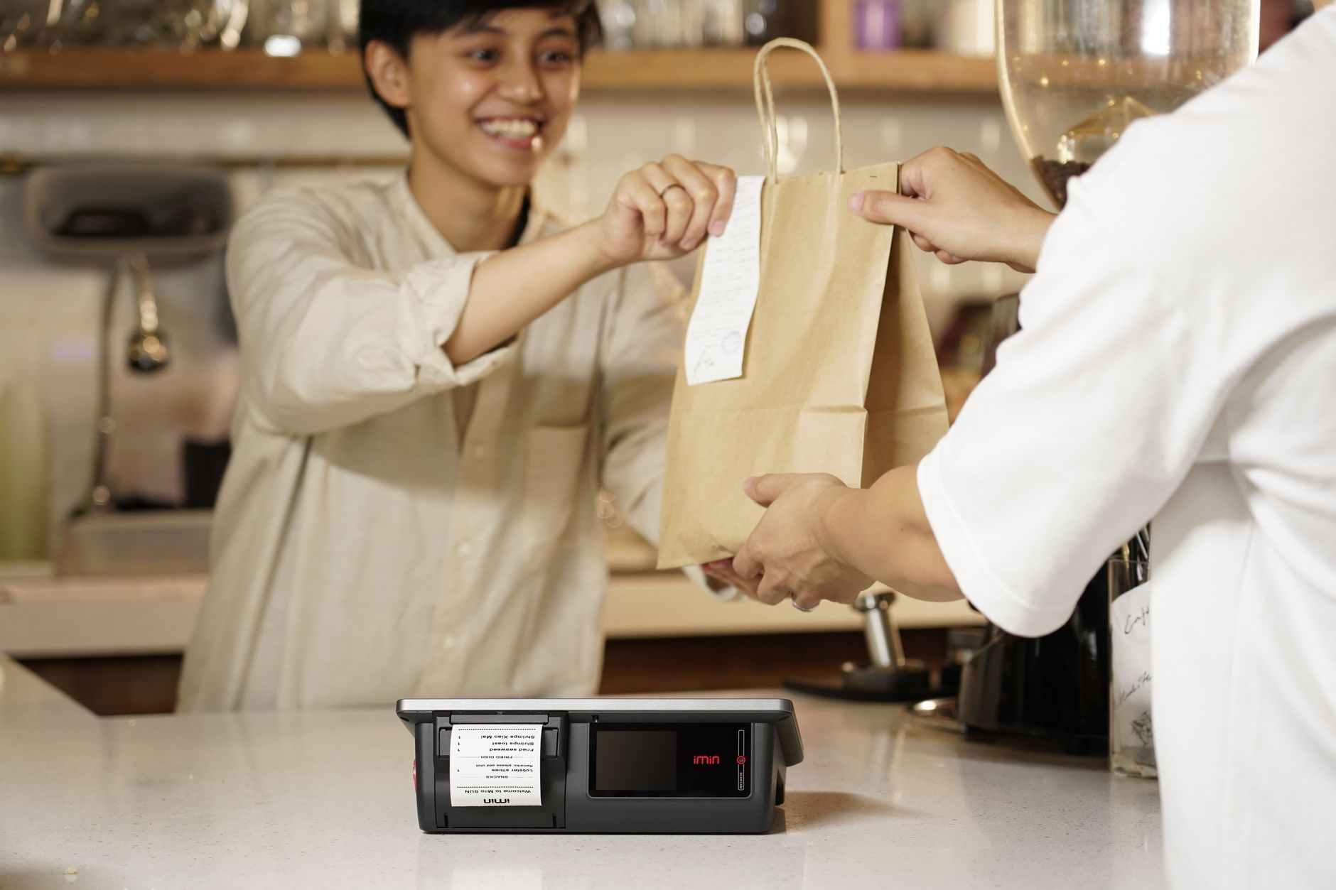 A smiling cashier handing a paper bag to a customer over the counter in a cozy cafe setting.