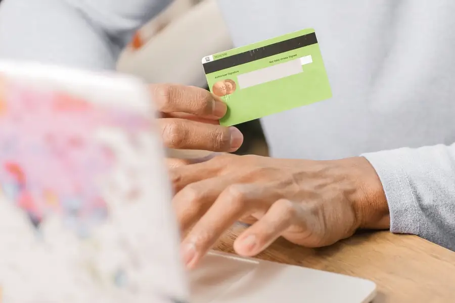 Mãos segurando um cartão de crédito verde diante de um notebook Mãos segurando um cartão de crédito verde diante de um notebook