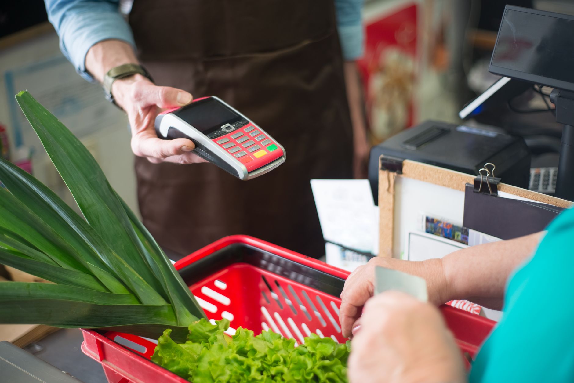 Pagar em euro ou reais no cartão internacional na Europa? A customer using a contactless payment method at a grocery store checkout with fresh produce.