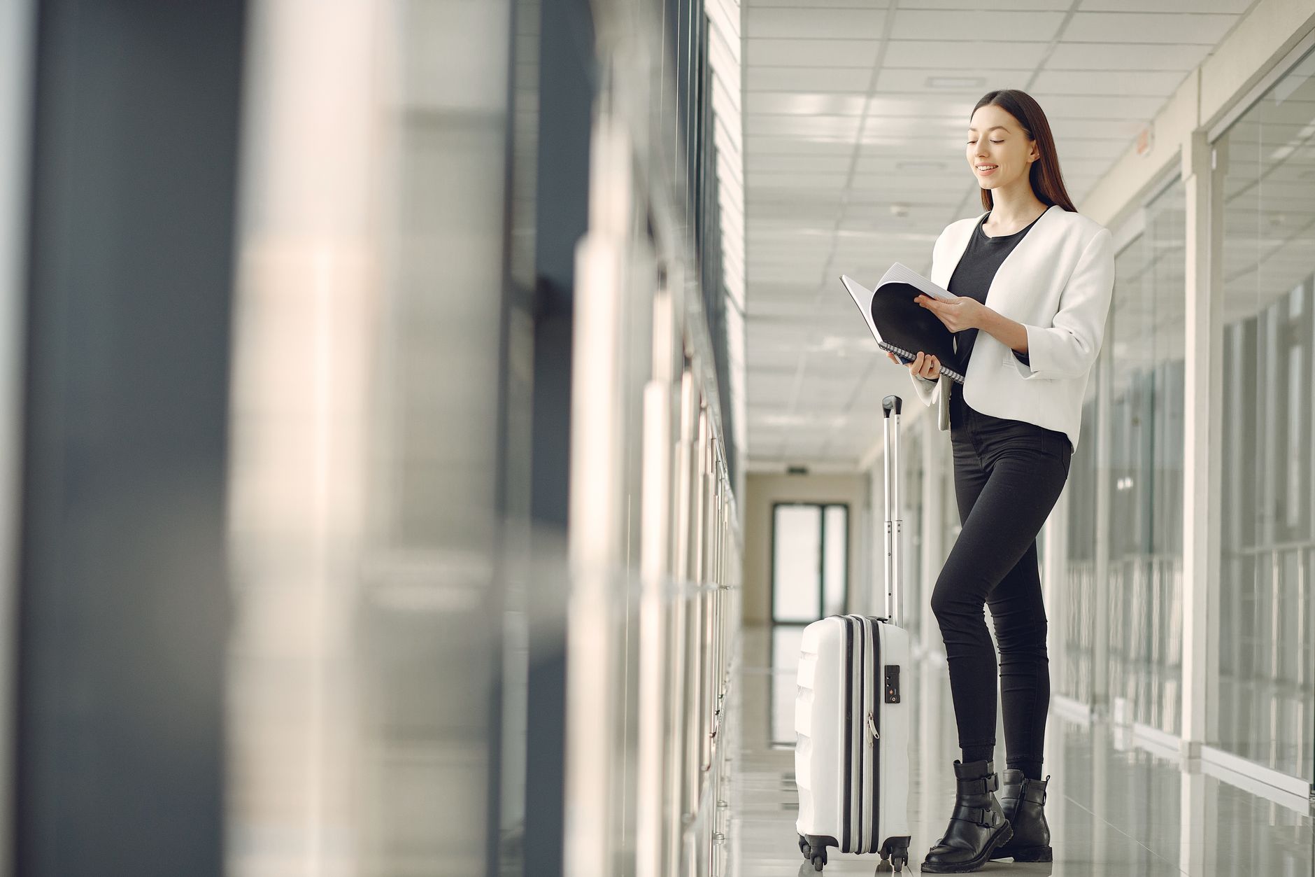 Qual é a multa por não declarar dinheiro em viagem internacional Full body of slim female businesswoman in formal clothes with suitcase standing in airport hallway and reading note in notebook while waiting for flight