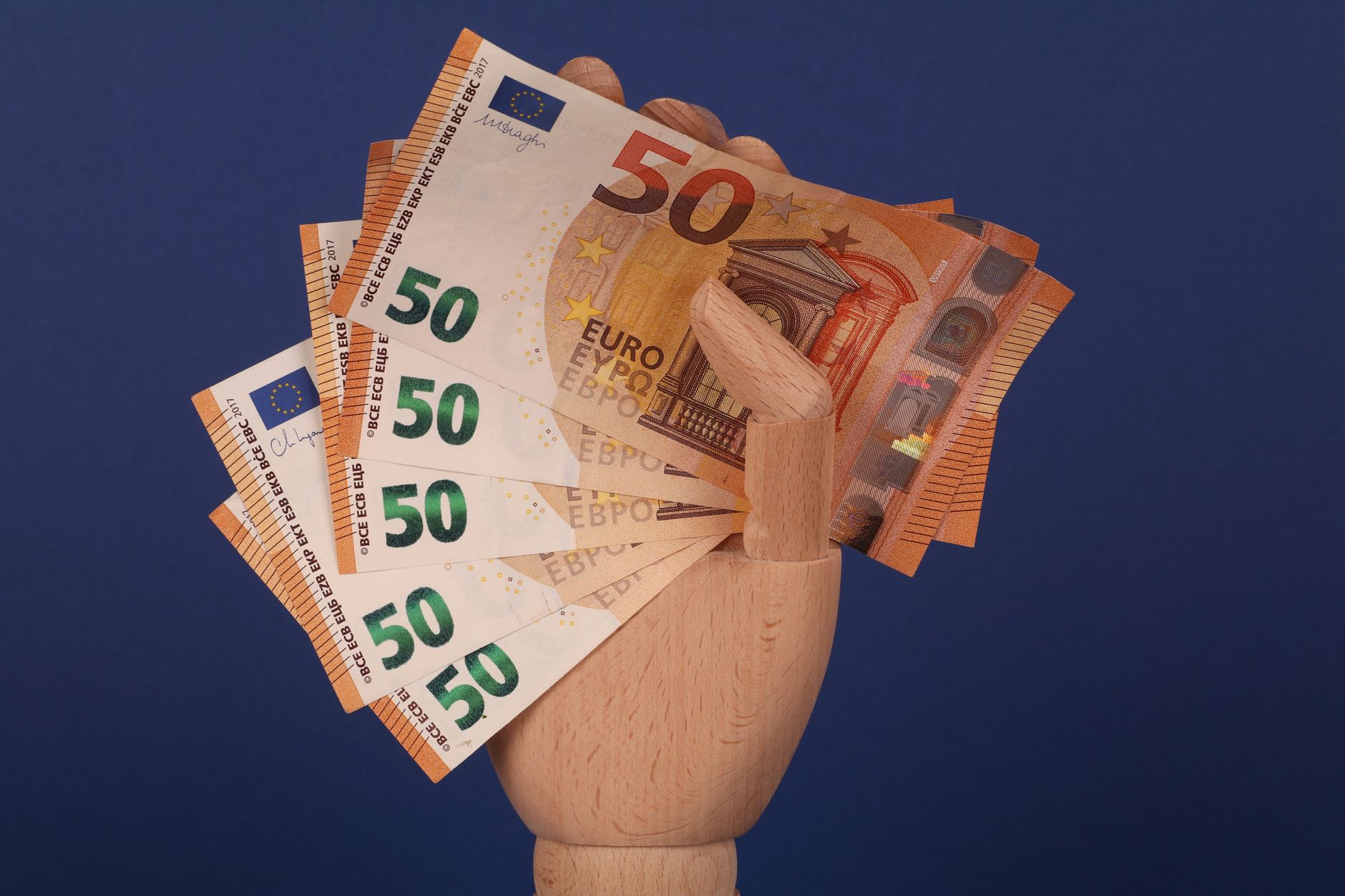 Quando o euro fica mais barato para brasileiros? A wooden mannequin hand grasping fifty euro banknotes with a blue backdrop.